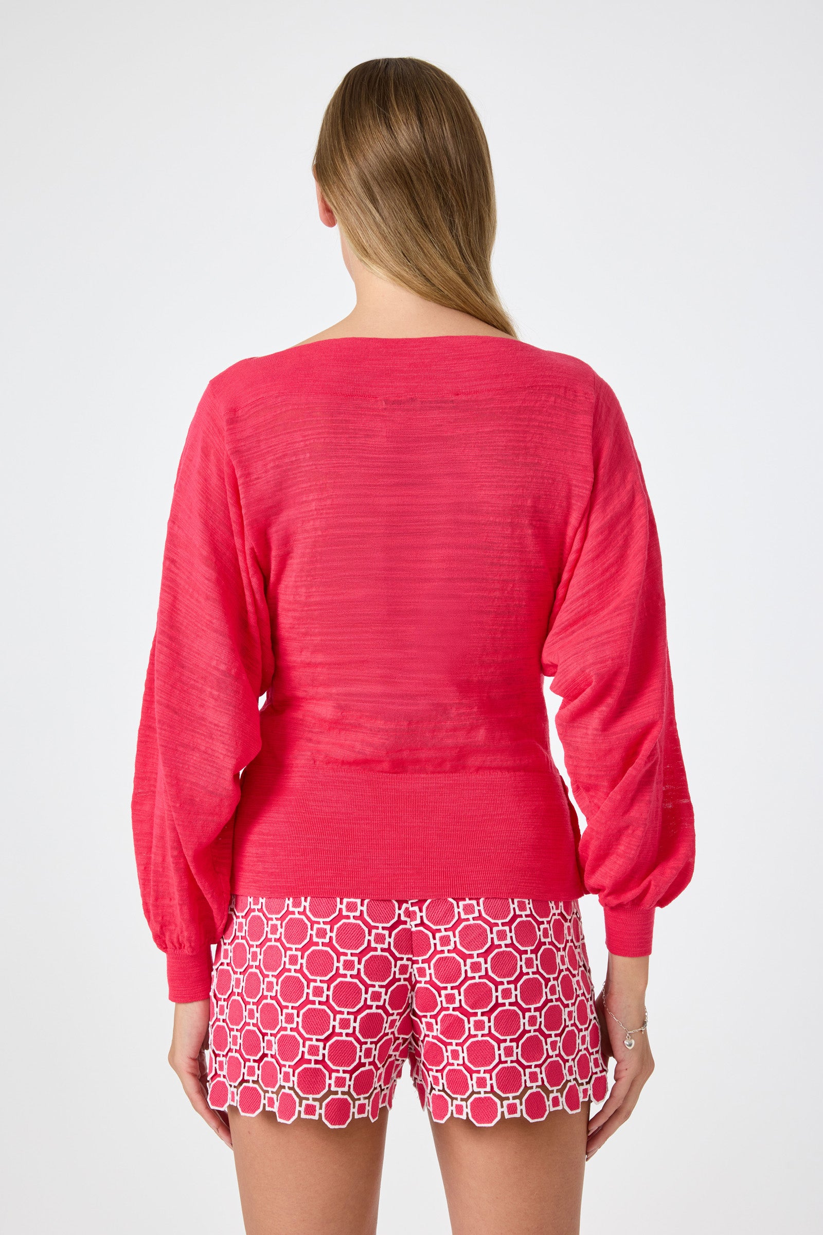 A woman faces away, wearing the Ramona 2 Boatneck Sweater in bright pink with long sleeves and patterned white-and-pink shorts. Her long hair drapes over one shoulder against a plain, light background.