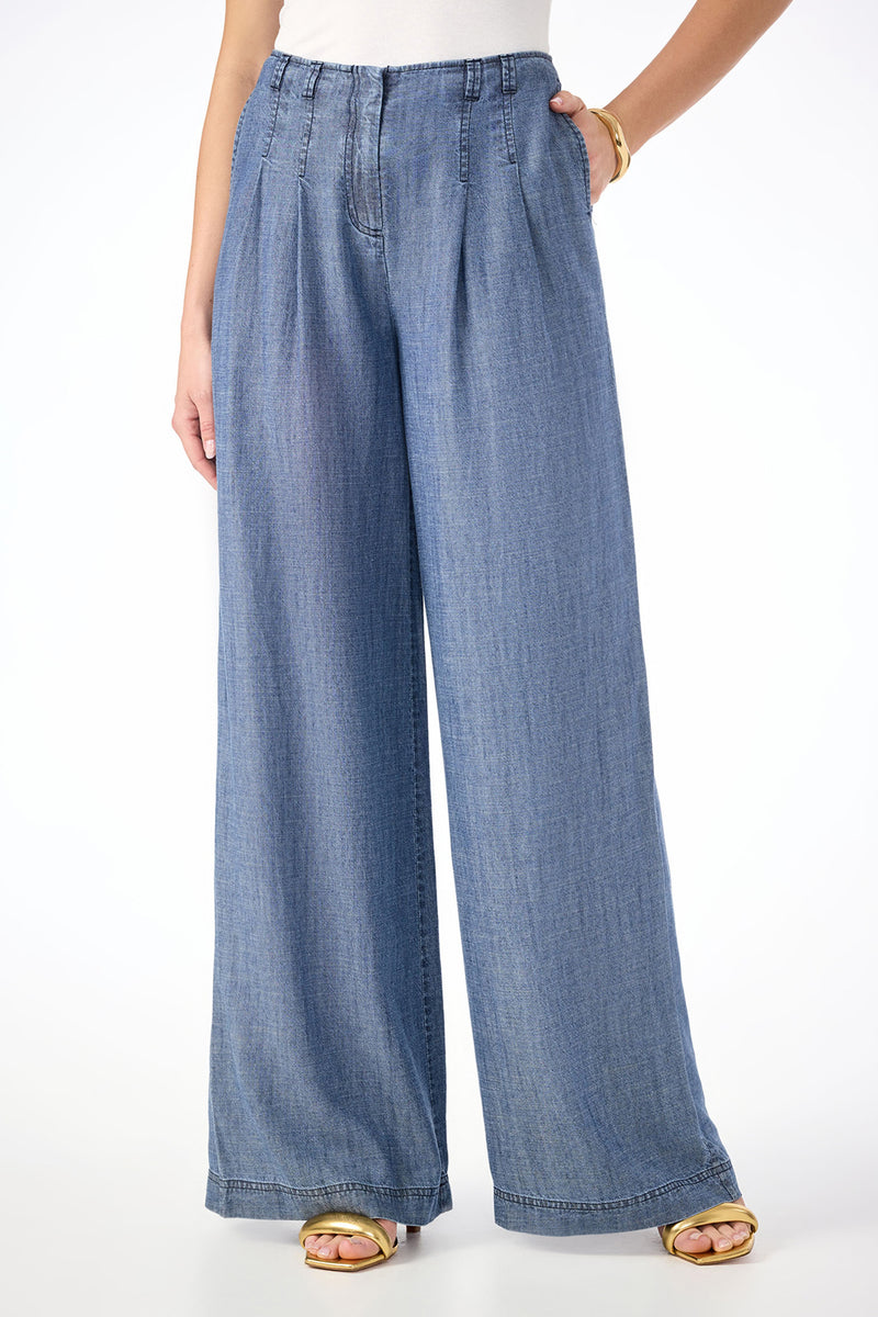A person stands against a white background wearing Dans Wide-Leg Chambray Pants in indigo, styled with gold sandals and a white top.