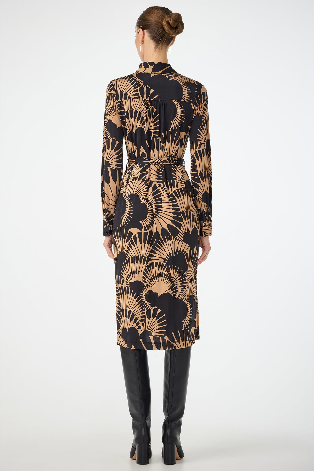 A woman faces away, wearing the Radio City Wrap Dress in Juniper Jersey—a black and tan patterned midi with long sleeves and a belted waist—paired with tall black boots. Her hair is in a neat bun against a plain white background.