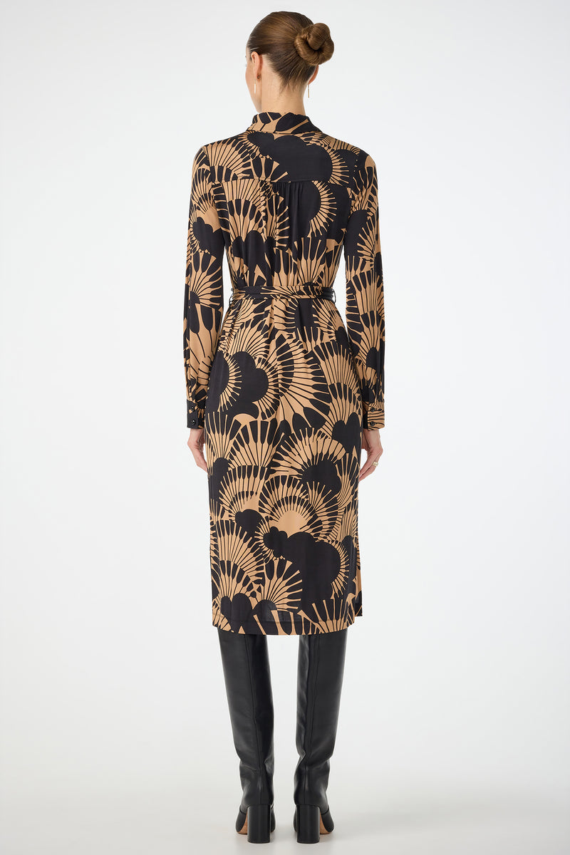 A woman faces away, wearing the Radio City Wrap Dress in Juniper Jersey—a black and tan patterned midi with long sleeves and a belted waist—paired with tall black boots. Her hair is in a neat bun against a plain white background.