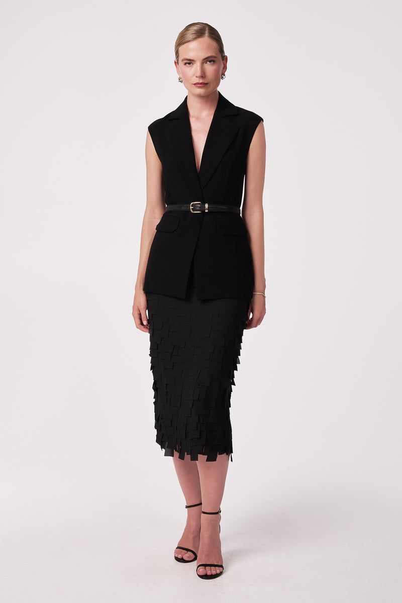 A woman stands against a plain background, wearing a sleeveless black belted blazer and the Dita Midi Pencil Skirt - Flutter Chiffon. Her hair is pulled back and she completes the look with black strappy heels.