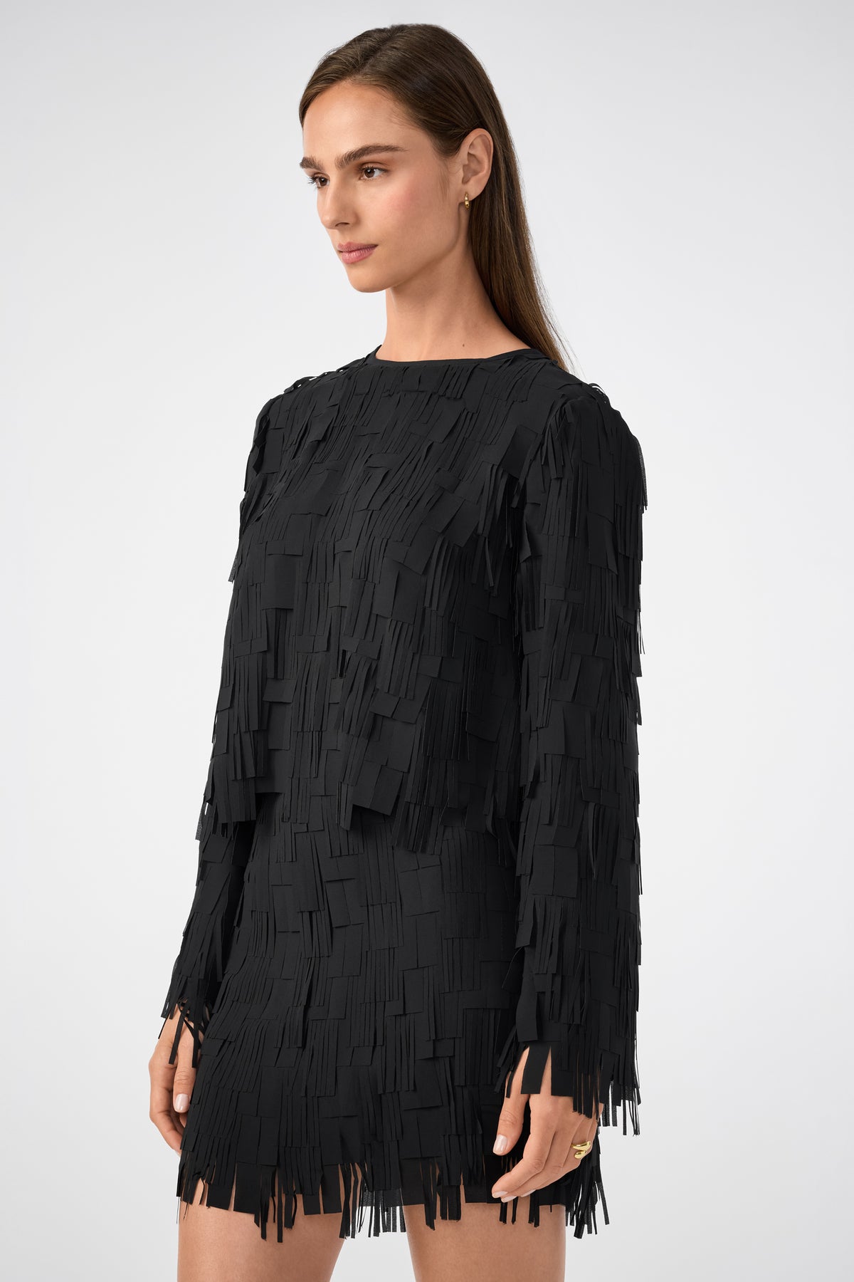 A woman in the Chinaberry Top, a long-sleeved black blouse with layered rectangular fringe details, stands against a plain light background, looking slightly to the side with a neutral expression.