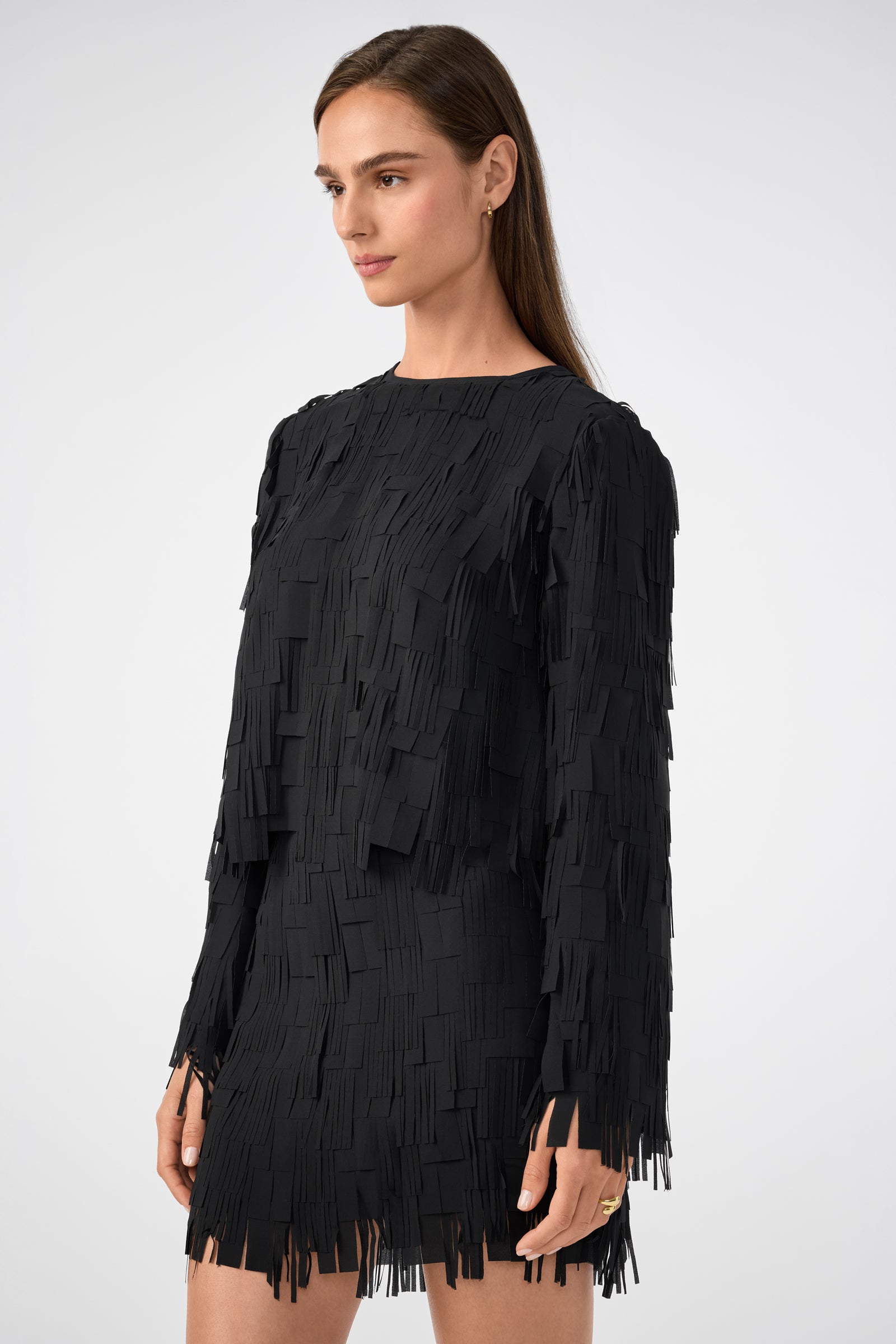 A woman in the Chinaberry Top, a long-sleeved black blouse with layered rectangular fringe details, stands against a plain light background, looking slightly to the side with a neutral expression.