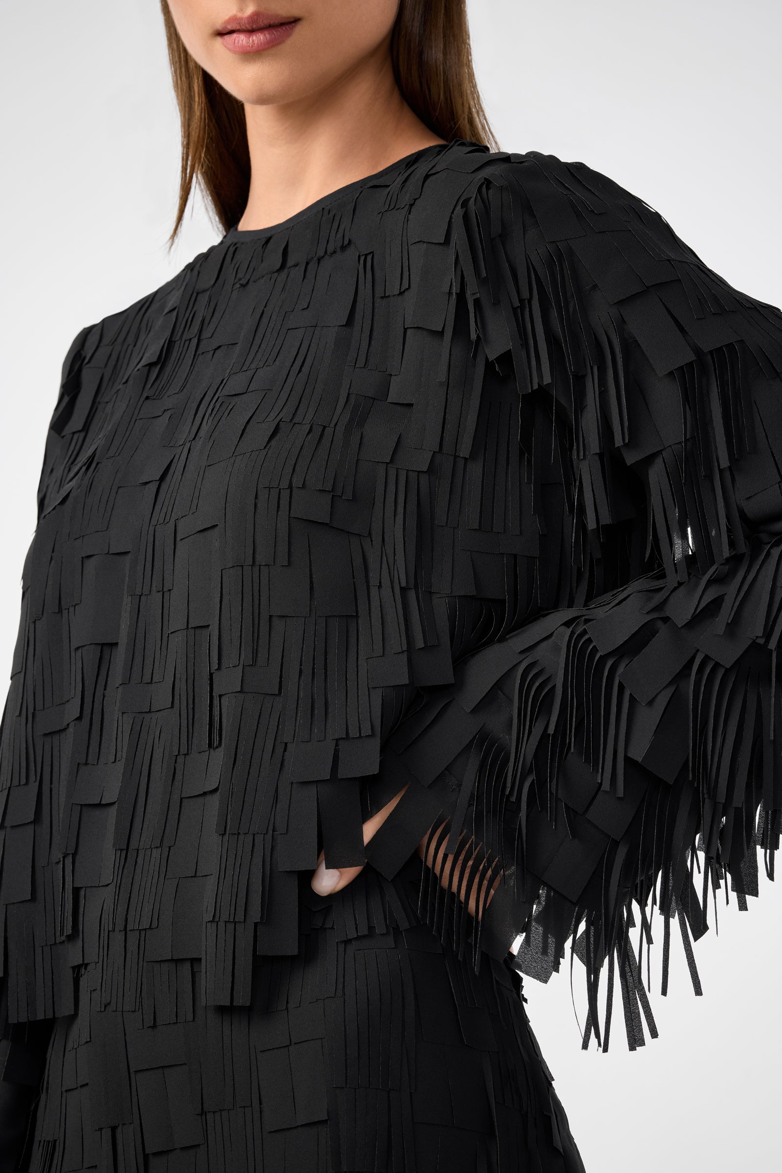 A woman wears the Chinaberry Top, a black long-sleeve style featuring layered, fringed rectangular fabric details. Only the lower half of her face is visible as she rests her hand on her hip.