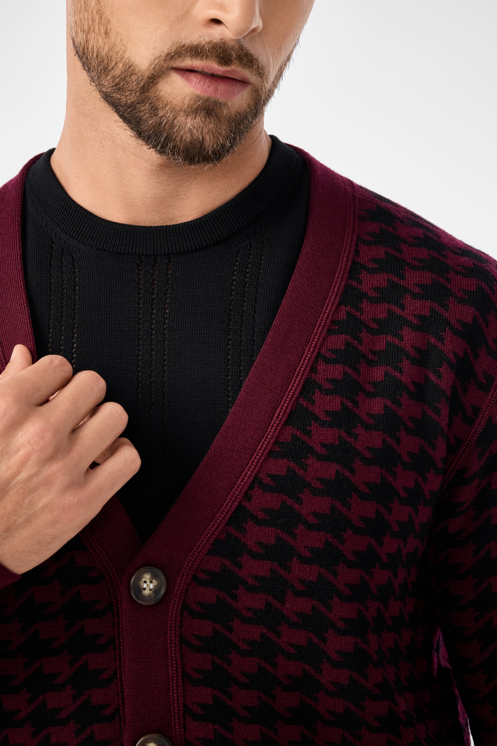 A man, partially visible from shoulders to mid-chest, touches the Alder Cardigan in burgundy and black houndstooth layered over a black sweater.