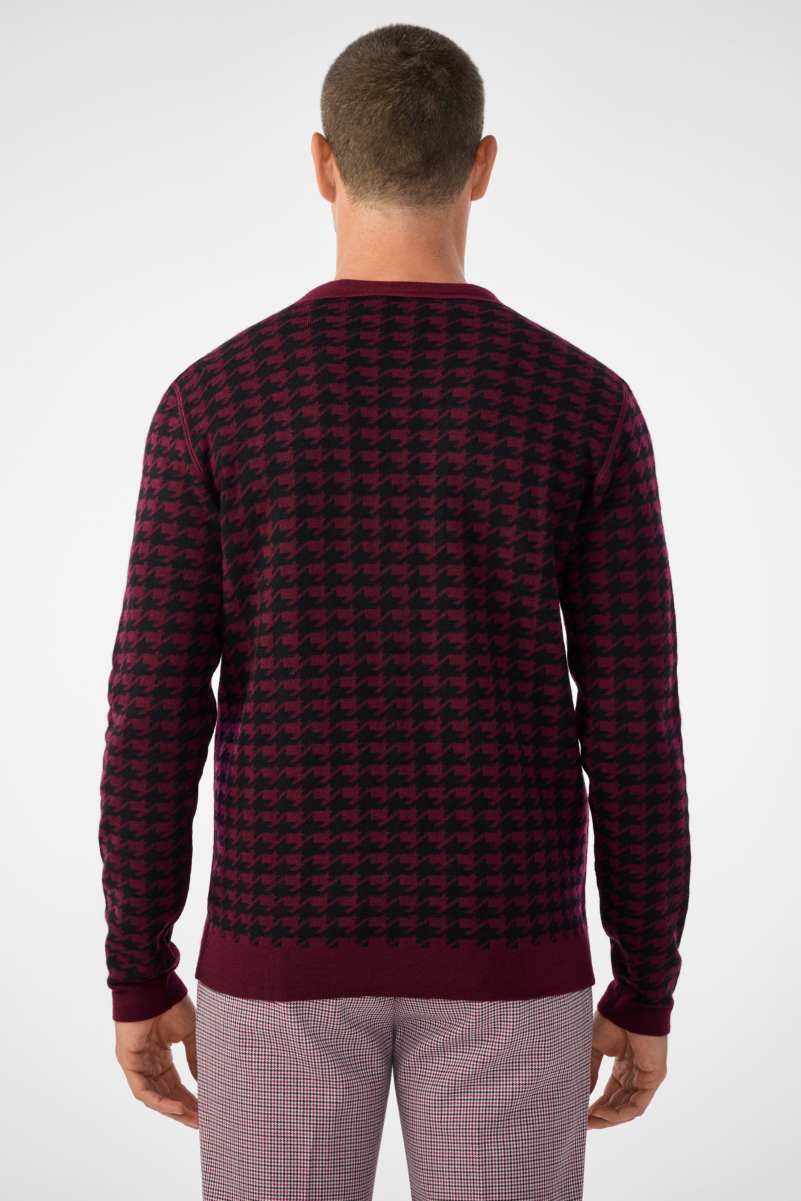 A man is seen from behind wearing the Alder Cardigan, featuring a burgundy and black houndstooth pattern, paired with light plaid pants against a plain light background.