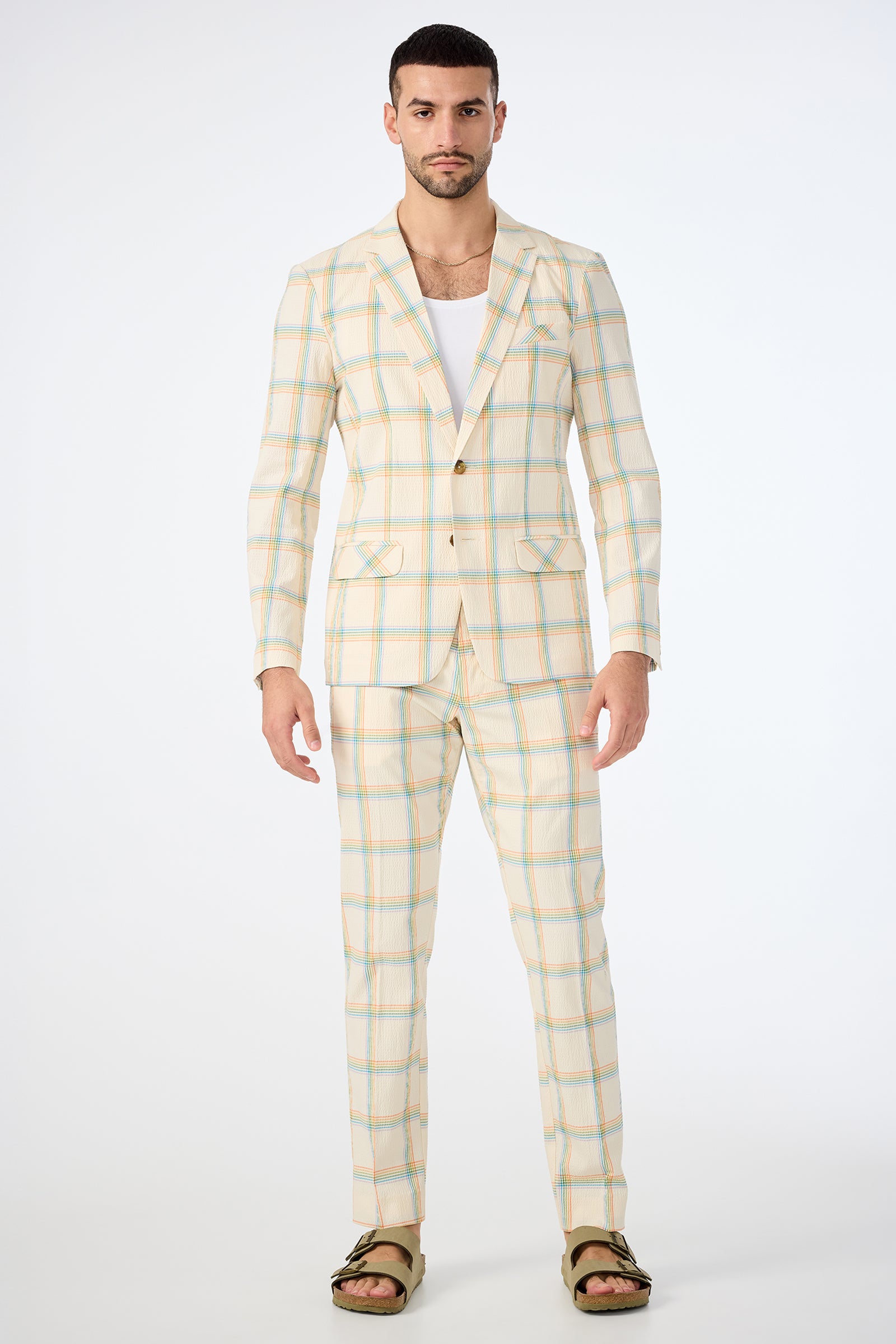 A man stands facing forward, wearing the Louie Blazer - Pride Plaid over a white undershirt and green sandals. The blazer features a light plaid pattern with pastel blue and orange stripes. The background is plain white.