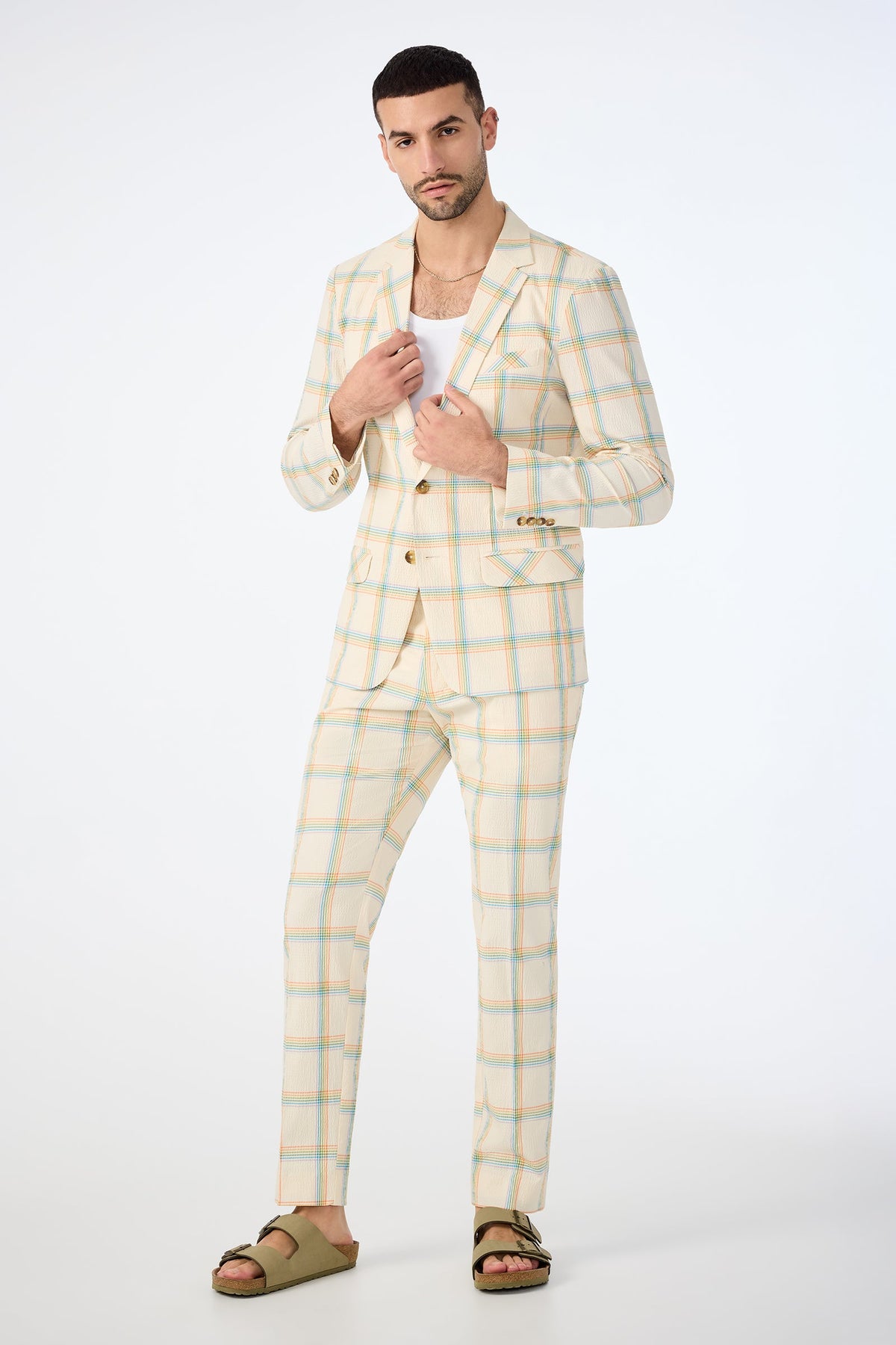 A man poses against a plain white background wearing the Clyde Slim Trouser in Pride Plaid, paired with a white shirt and olive green sandals. He has short dark hair and a trimmed beard.