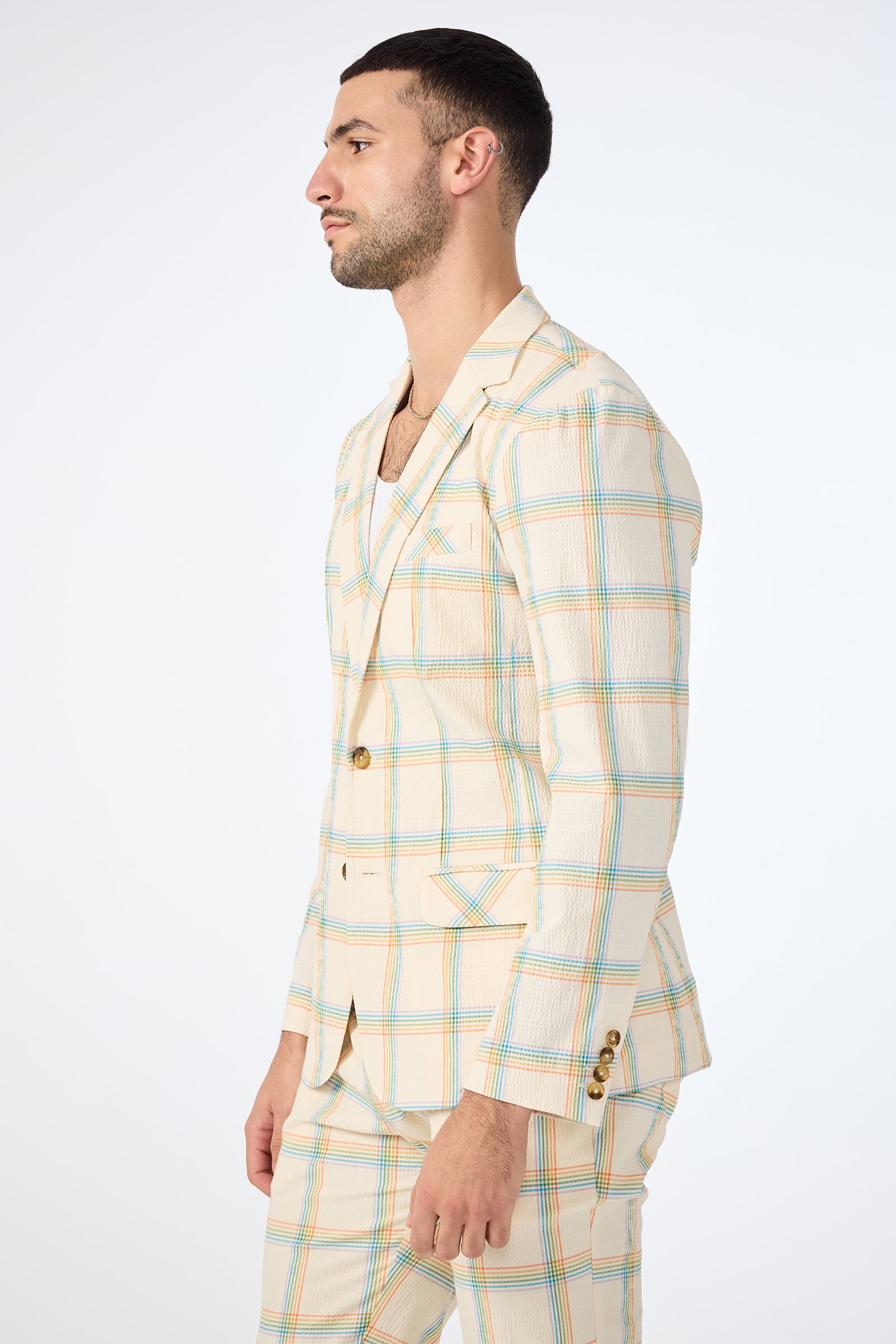 A man stands in profile against a plain white background, wearing the Louie Blazer - Pride Plaid, a cream-colored suit featuring blue, orange, and yellow plaid accents.