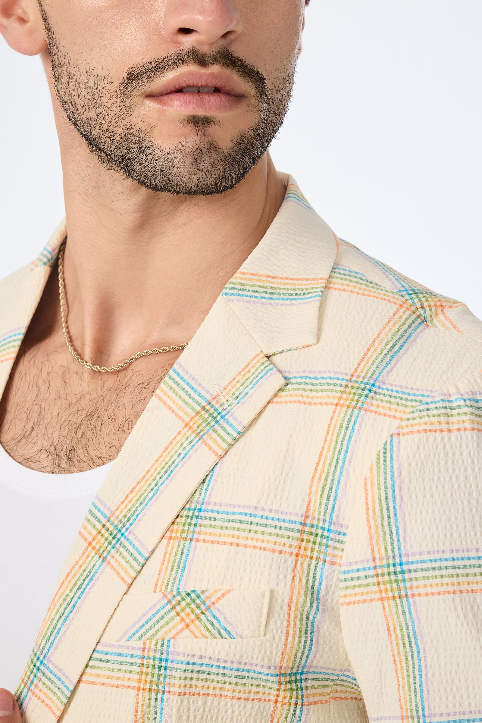 A man wears the Louie Blazer - Pride Plaid, a cream blazer with a pastel plaid pattern, paired with a white shirt and gold chain. The photo focuses on his lower face, neck, and upper chest.