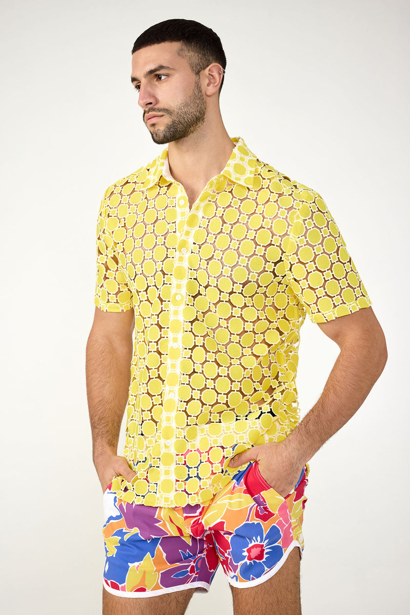 A man stands with hands in pockets, looking to the side against a plain background, wearing the Long Beach Short-Sleeve Shirt—a sheer yellow button-up with a circular pattern—paired with colorful floral shorts.