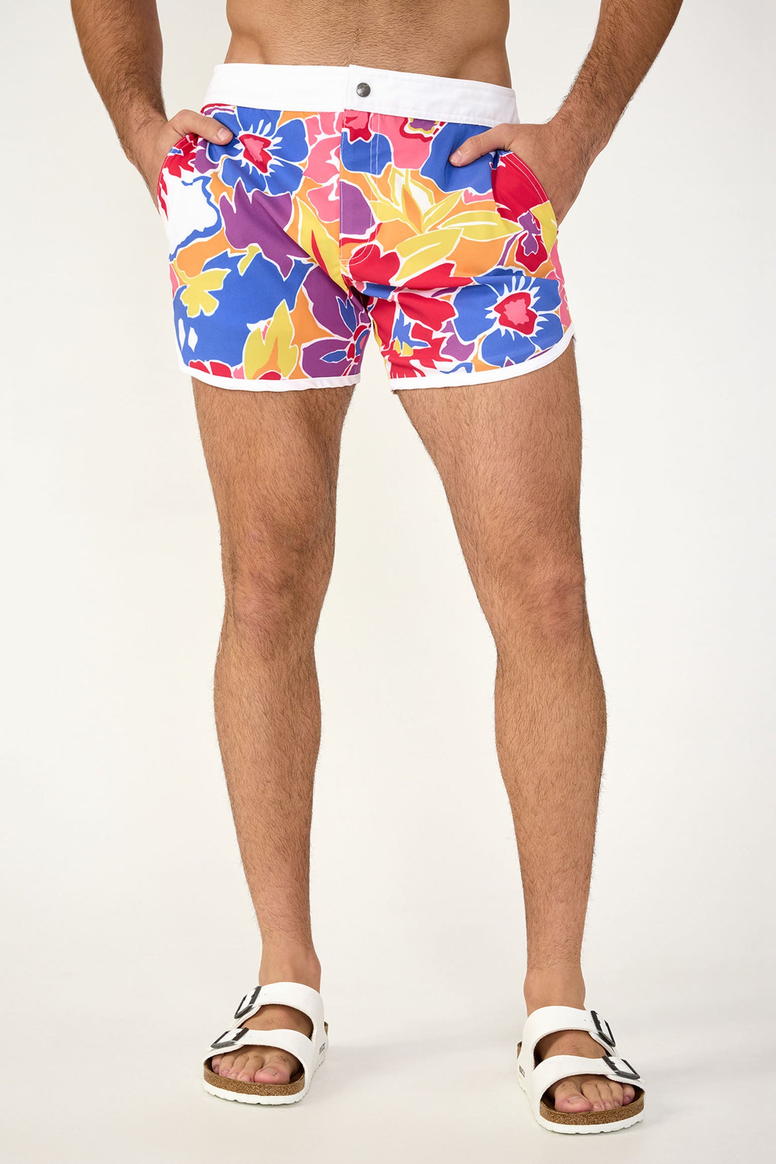 A person wearing Surfside Board Short in Soie Paradis, featuring colorful floral patterns, with white sandals stands against a plain light background. Only their legs and lower torso are visible.