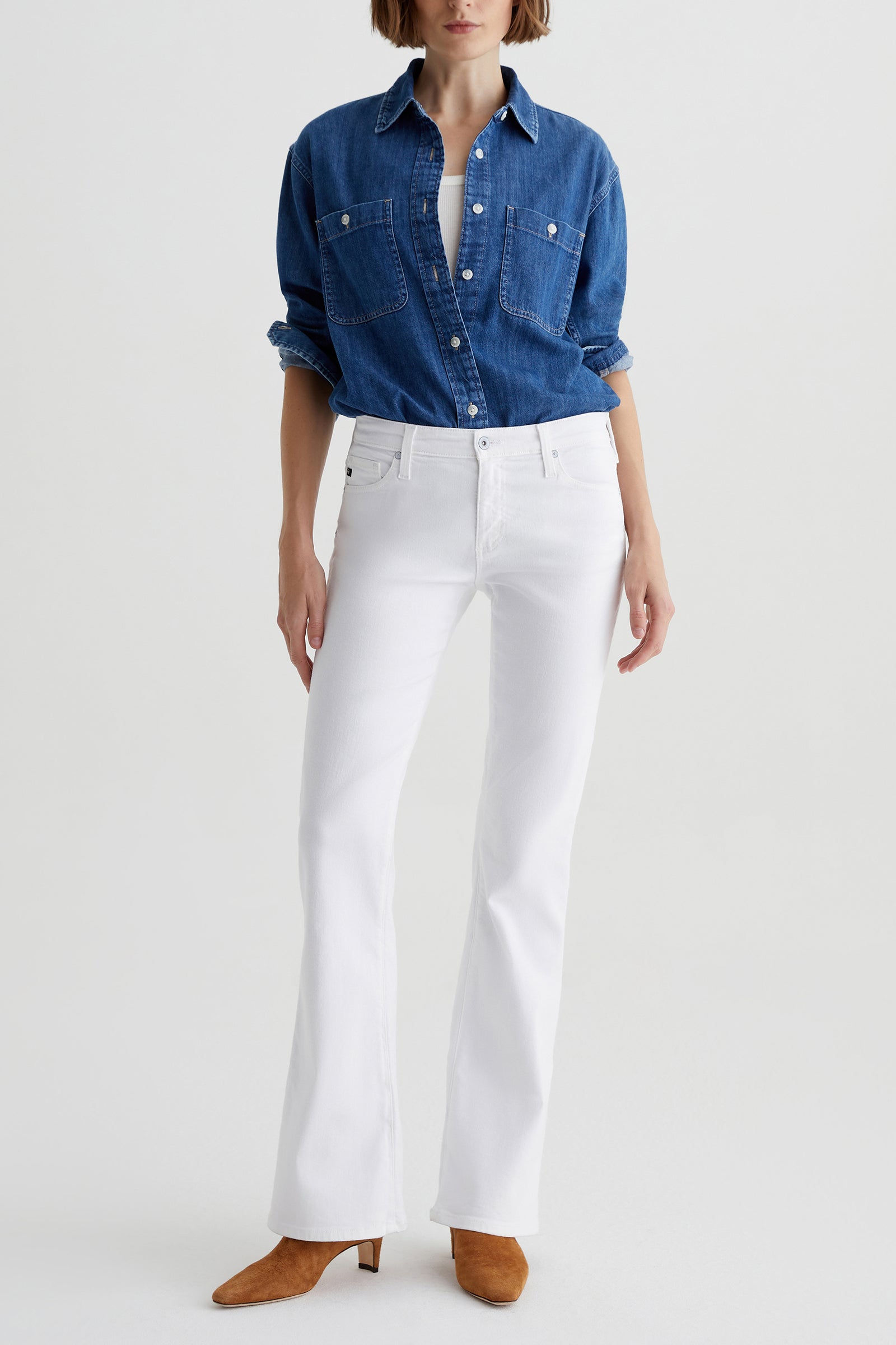A woman models the AG Angel Low-Rise Bootcut Jean in white, paired with a blue denim shirt and brown heeled boots, standing against a plain white background. Her hands rest at her sides and her head is partially out of frame.