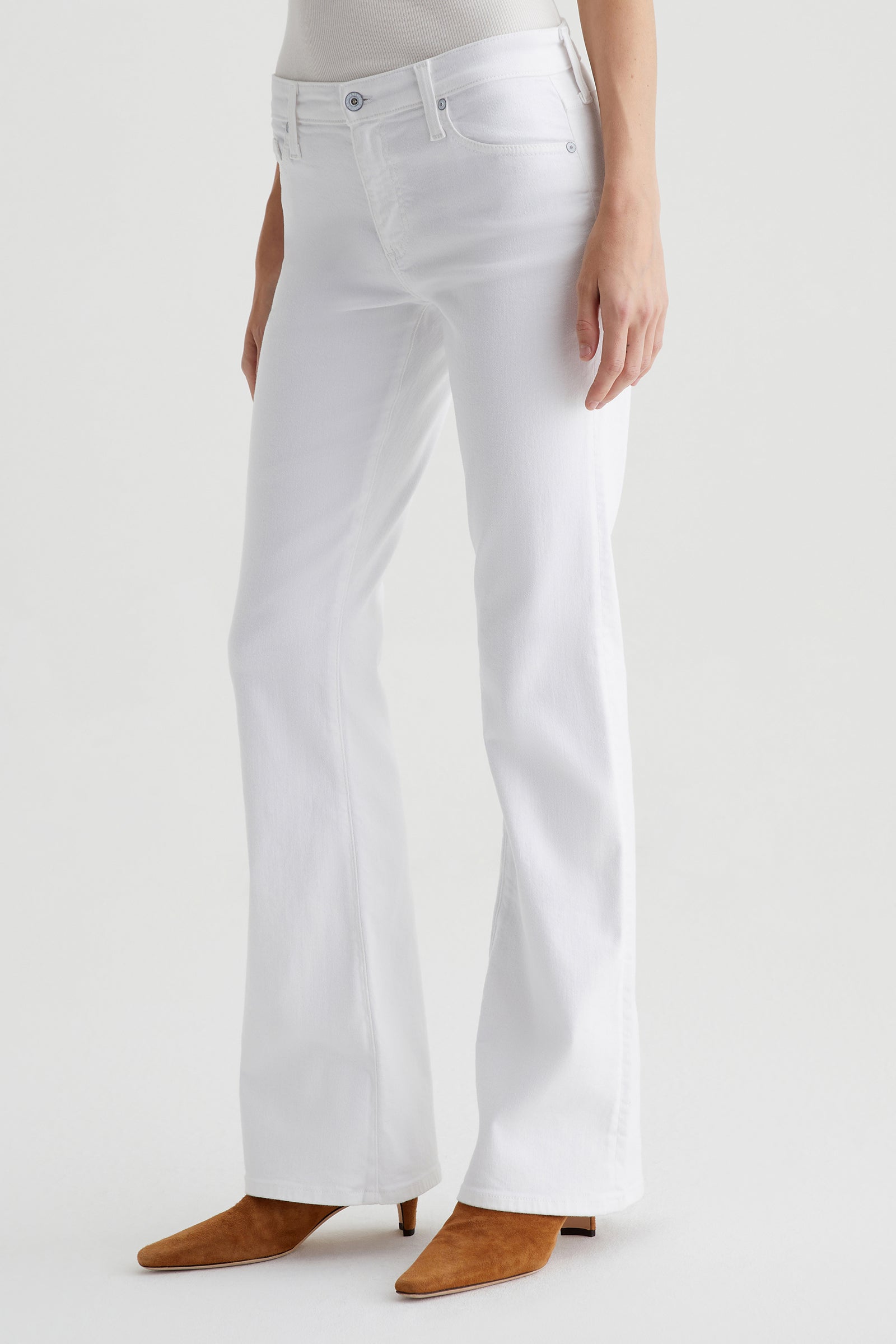 Wearing AG Angel Low-Rise Bootcut Jean in white and tan suede pointed-toe boots, a person stands against a plain white background. The image is cropped from the shoulders down.