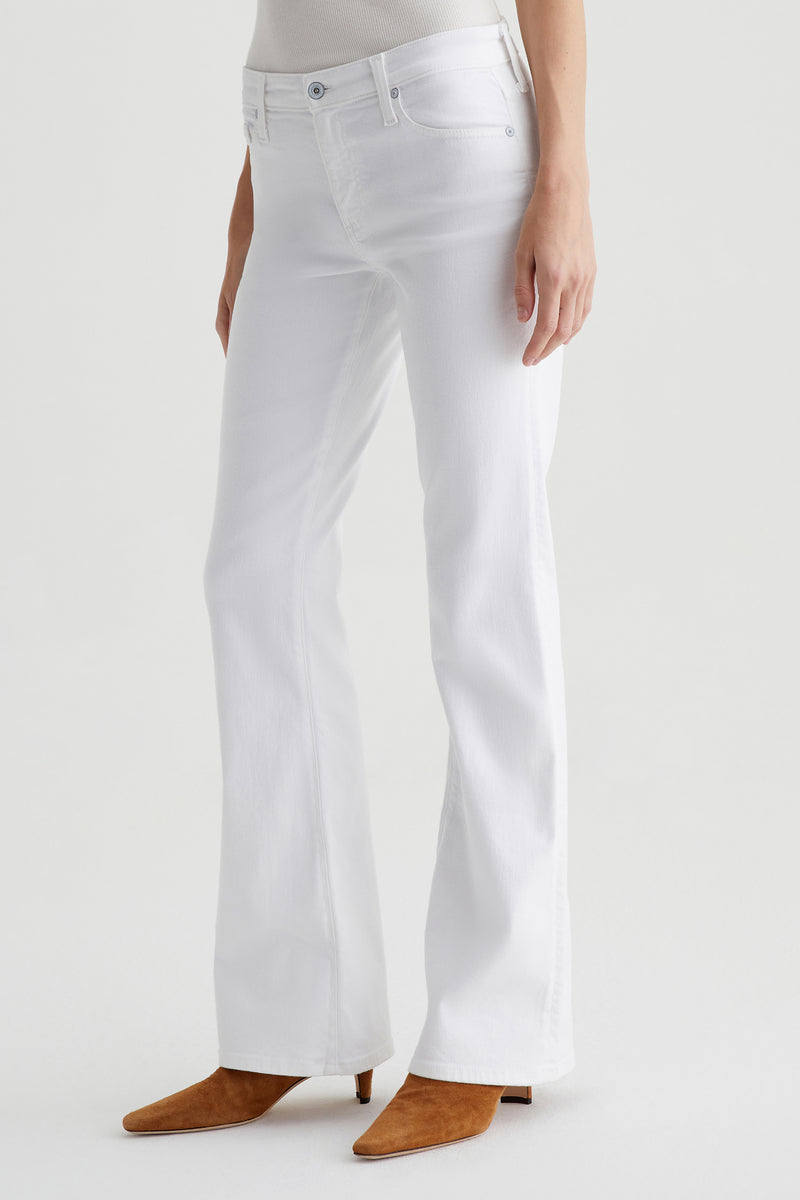 Wearing AG Angel Low-Rise Bootcut Jean in white and tan suede pointed-toe boots, a person stands against a plain white background. The image is cropped from the shoulders down.