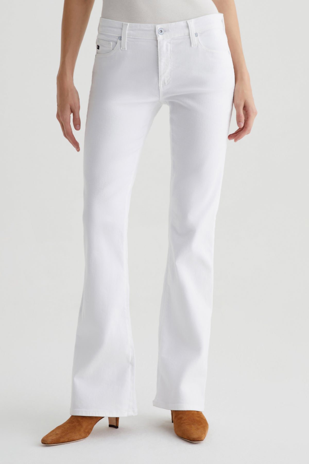 A person wearing AG Angel Low-Rise Bootcut Jean in white and tan suede pointed-toe boots stands against a plain light background, with their upper body partially out of frame.