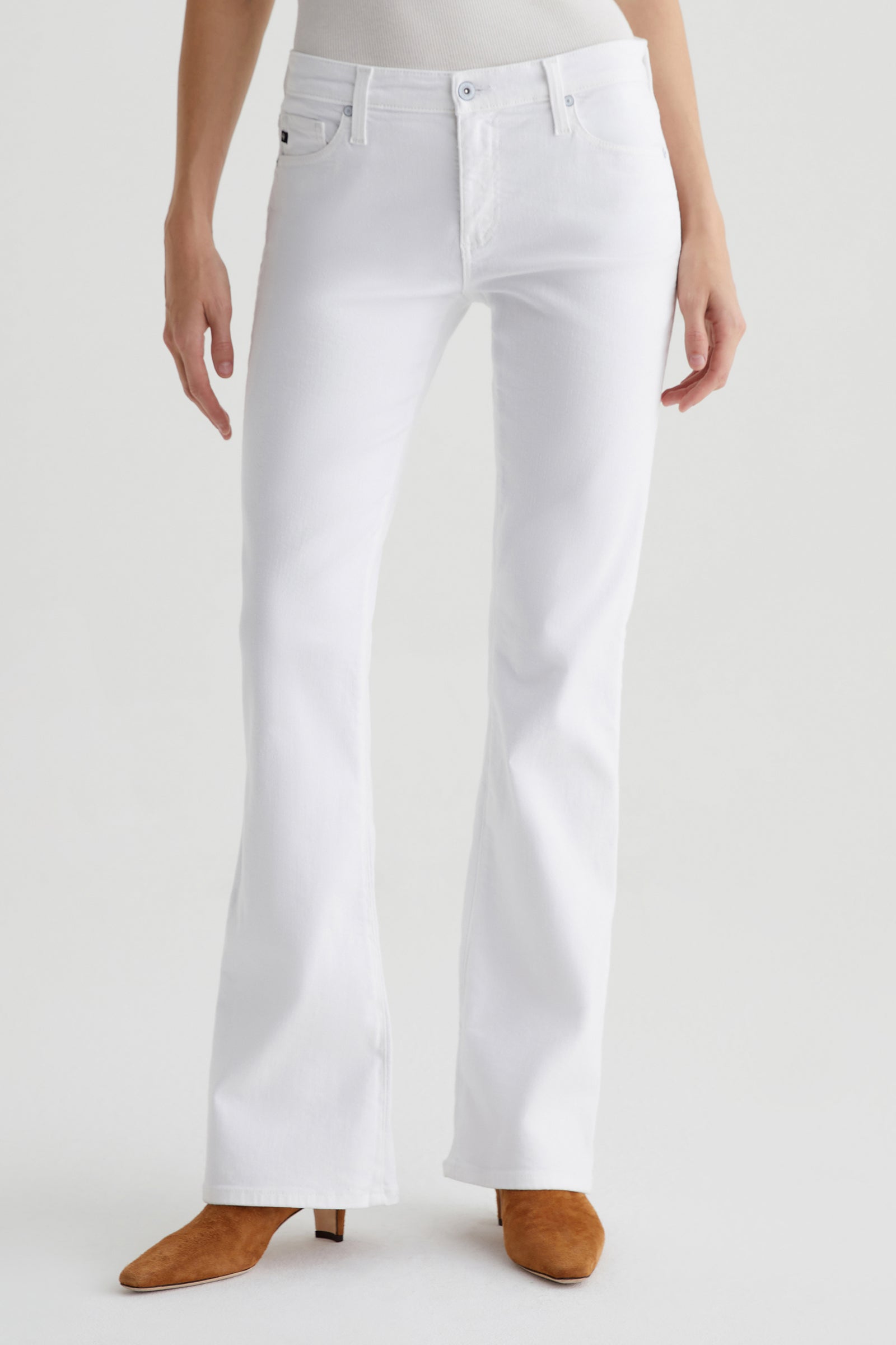 A person wearing AG Angel Low-Rise Bootcut Jean in white and tan suede pointed-toe boots stands against a plain light background, with their upper body partially out of frame.