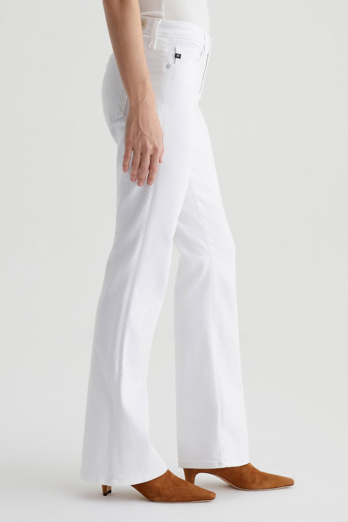 A person models the AG Angel Low-Rise Bootcut Jean in white with brown pointed-toe heeled boots, standing against a plain white background. Only the lower body is visible.