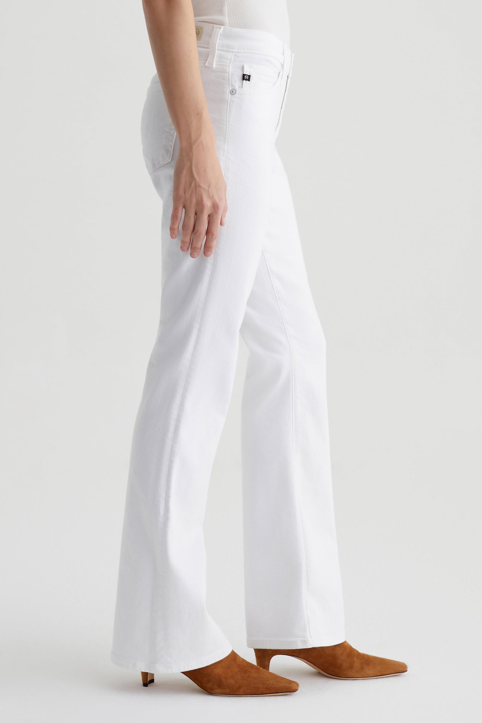 A person models the AG Angel Low-Rise Bootcut Jean in white with brown pointed-toe heeled boots, standing against a plain white background. Only the lower body is visible.