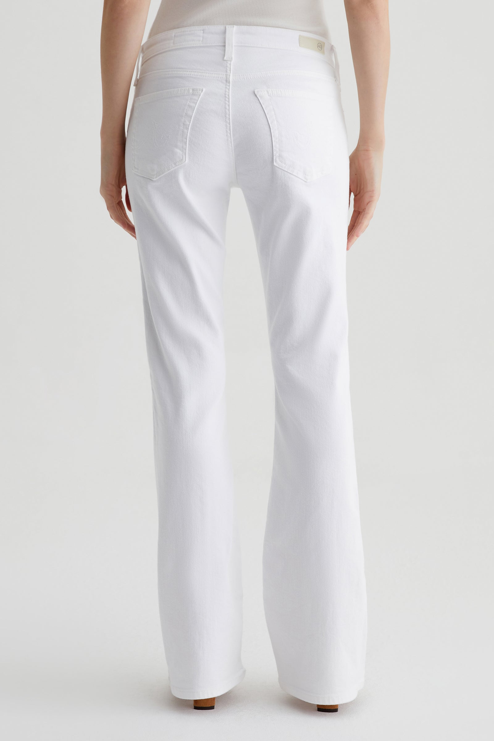 A person stands facing away from the camera, wearing the AG Angel Low-Rise Bootcut Jean in white and a light-colored top, set against a plain white background.