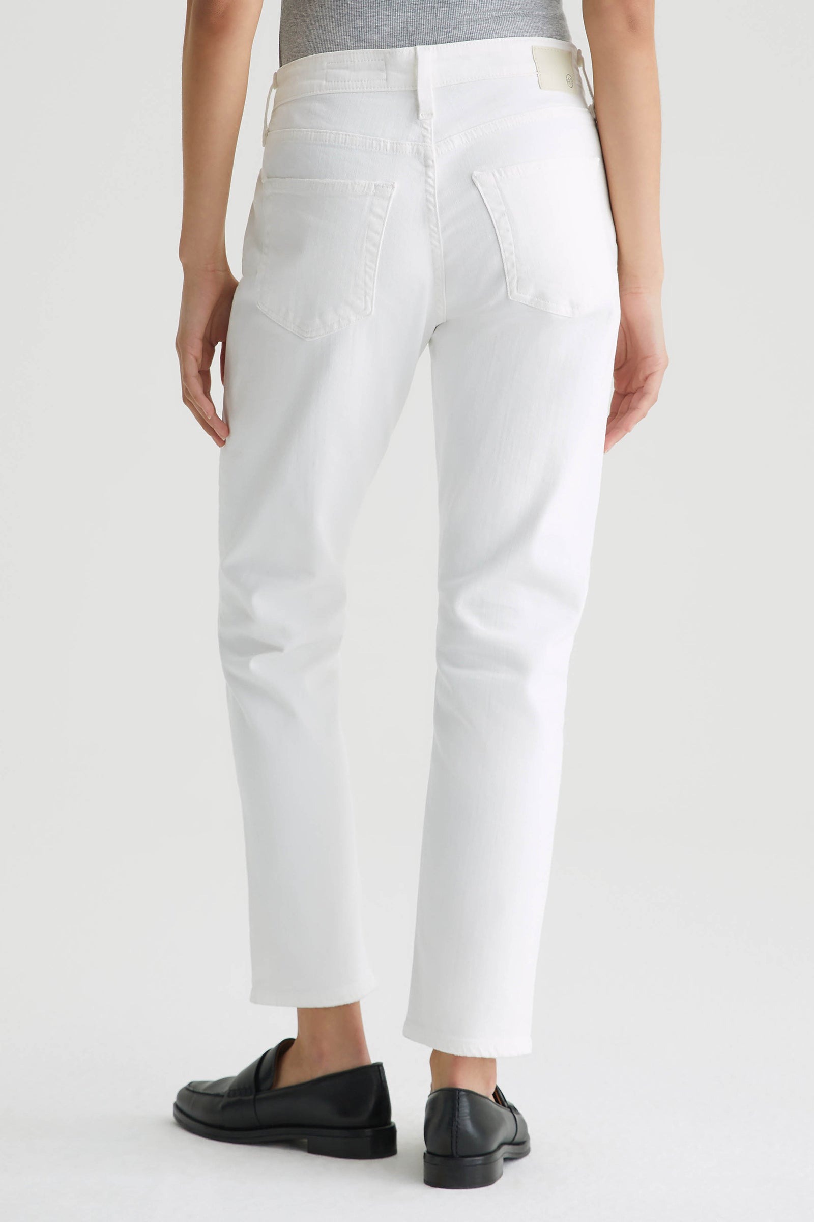 Person stands facing away in a gray top tucked into AG Mid-Rise Slouchy Ex Boyfriend Jean - White with black loafers; the image highlights the relaxed fit and back view of the jeans against a plain background.