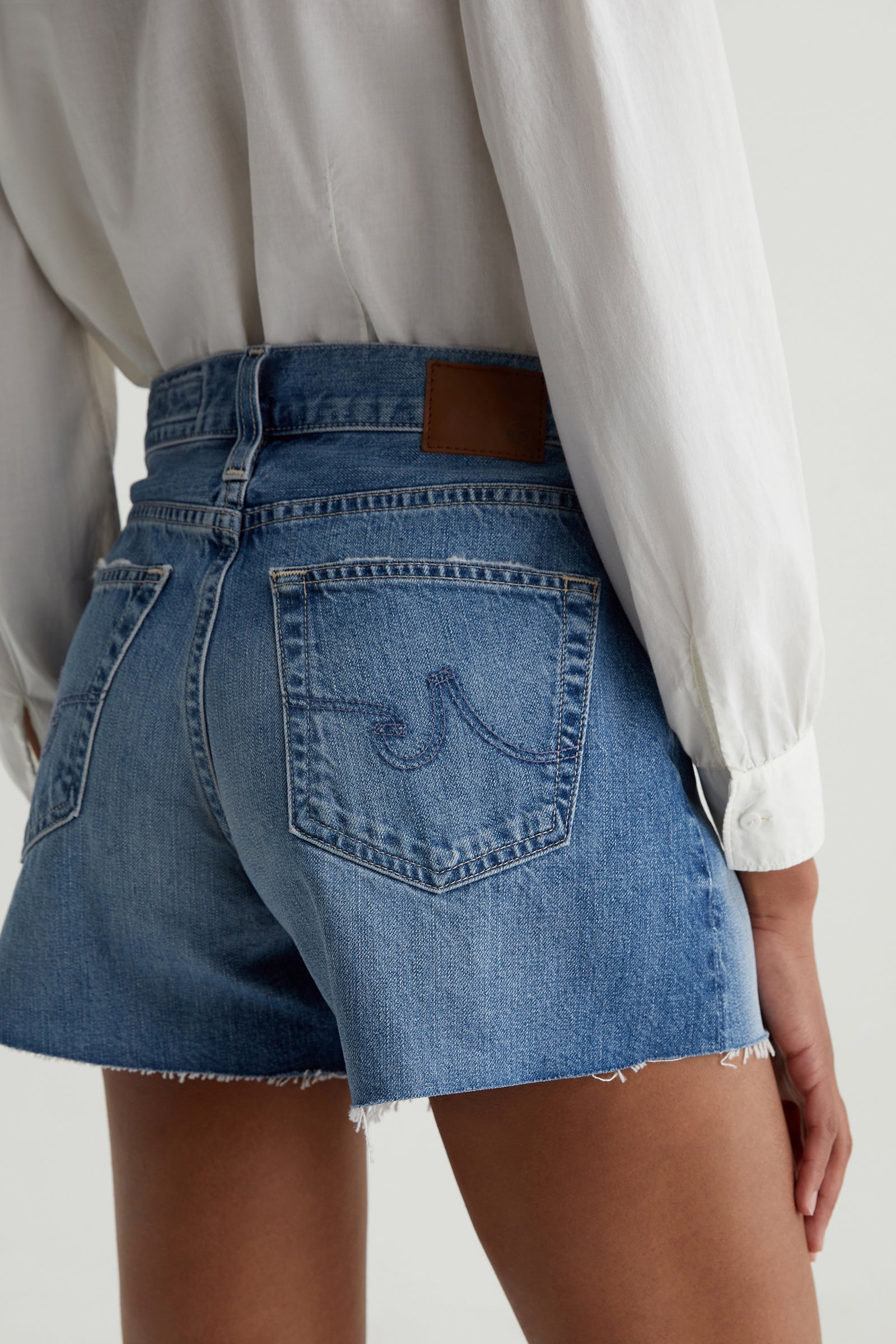 A person wearing a white long-sleeve shirt and the AG Halle Denim Short - Blue, featuring high-waisted fit and frayed hems, is shown from the back against a plain background.