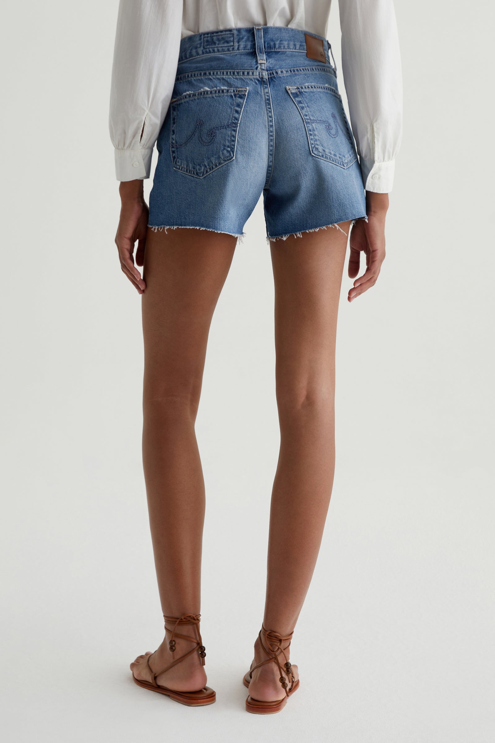 A person stands facing away against a plain light background, wearing a white long-sleeve blouse, brown strappy flat sandals, and the AG Halle Denim Short - Blue with frayed hems.