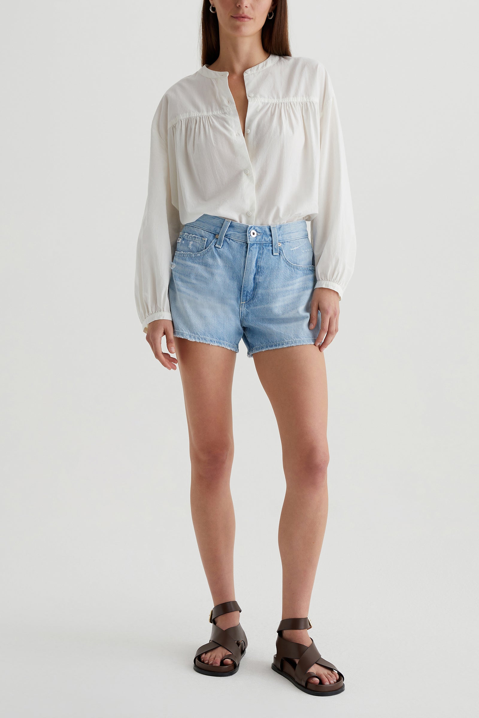 A woman stands facing forward against a plain white background, wearing a loose white long-sleeve blouse, dark brown sandals, and the AG Halle Denim Short in Light Blue. Her hair is down.