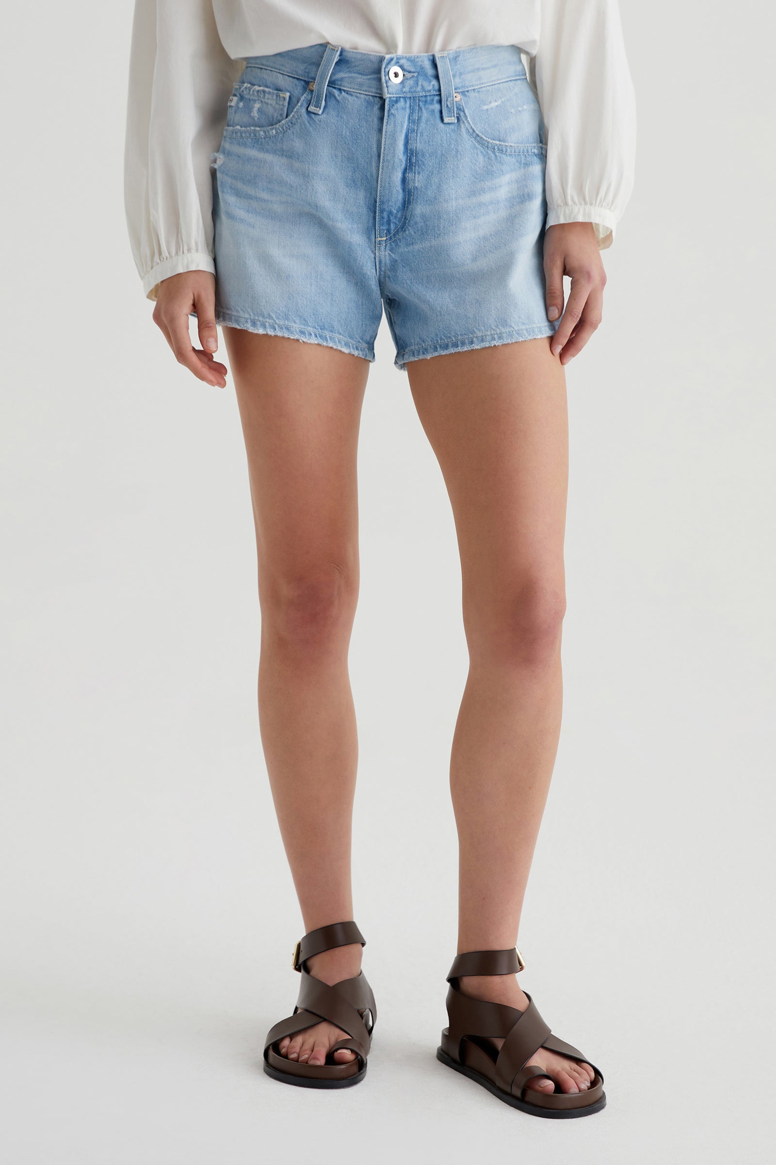 A person in the AG Halle Denim Short - Light Blue, paired with a white long-sleeve top and brown strappy sandals, stands against a plain light background. The photo is cropped at the shoulders.