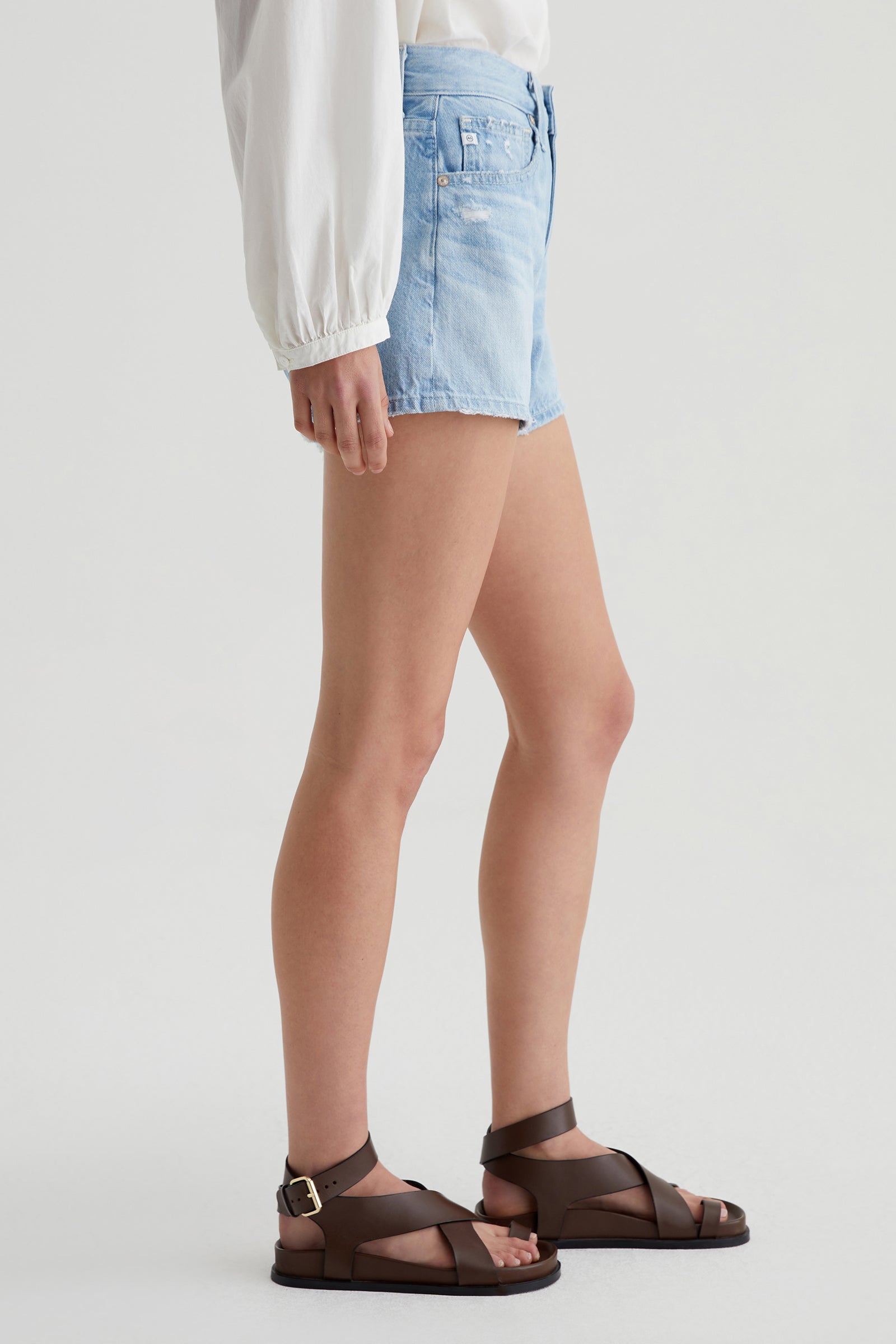 A person stands sideways against a plain light background, wearing a white long-sleeve blouse, brown strappy sandals, and AG Halle Denim Short in Light Blue.