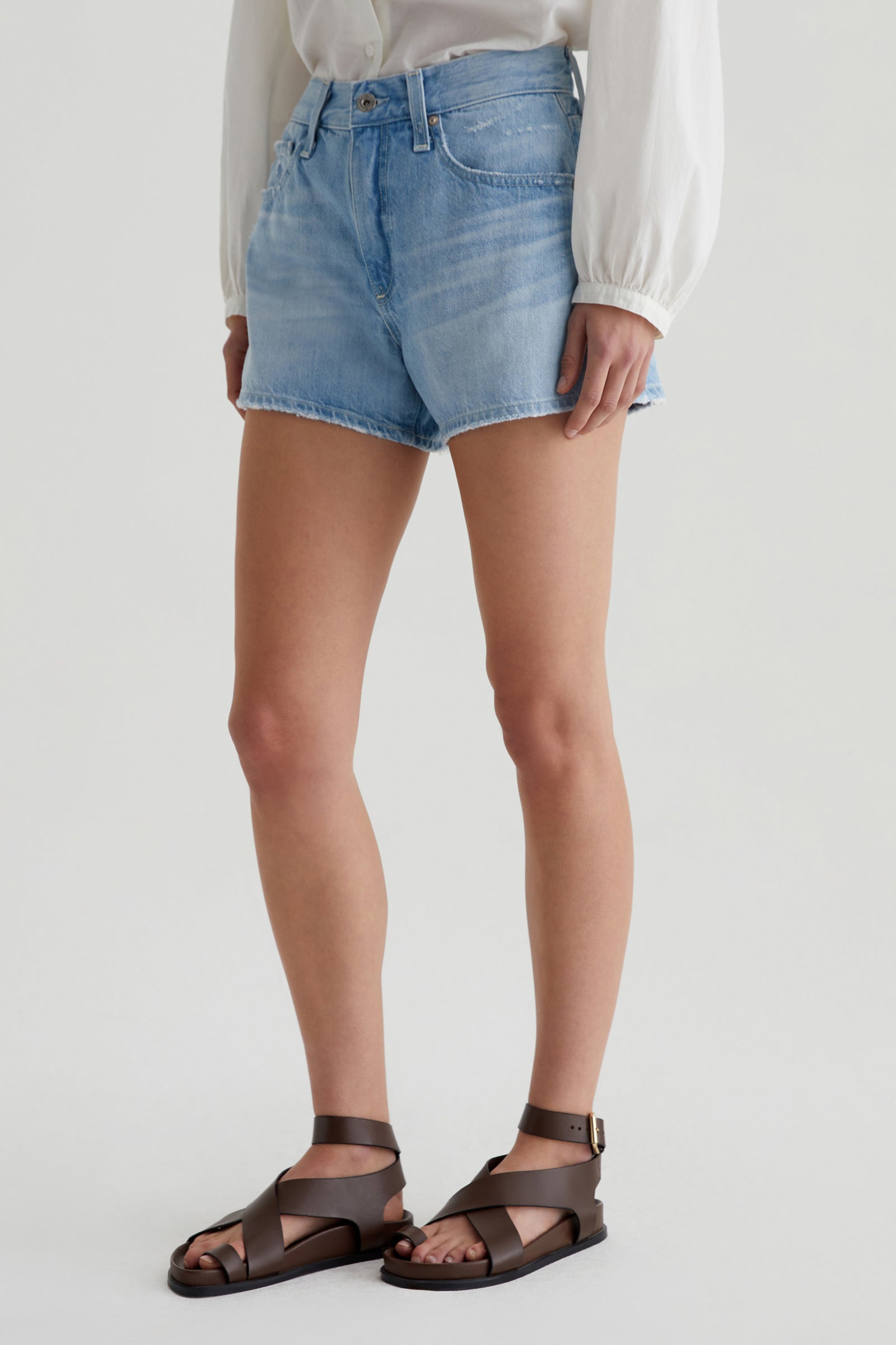Someone wearing AG Halle Denim Short - Light Blue, a white long-sleeve blouse, and brown leather sandals stands against a plain light background.