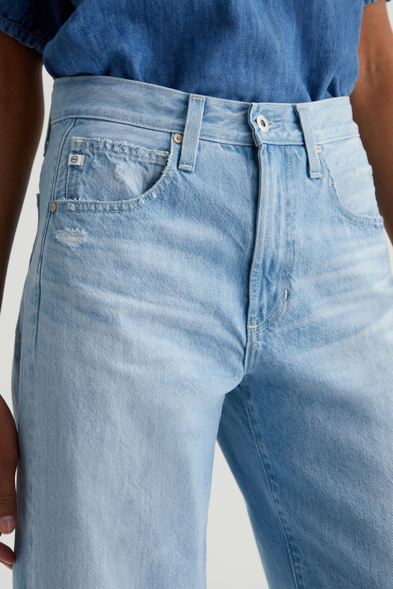 Close-up of a person wearing the AG Hattie Crop Jean - Light Blue with high waist and slight distressing near the front pocket, paired with a tucked-in blue denim shirt.