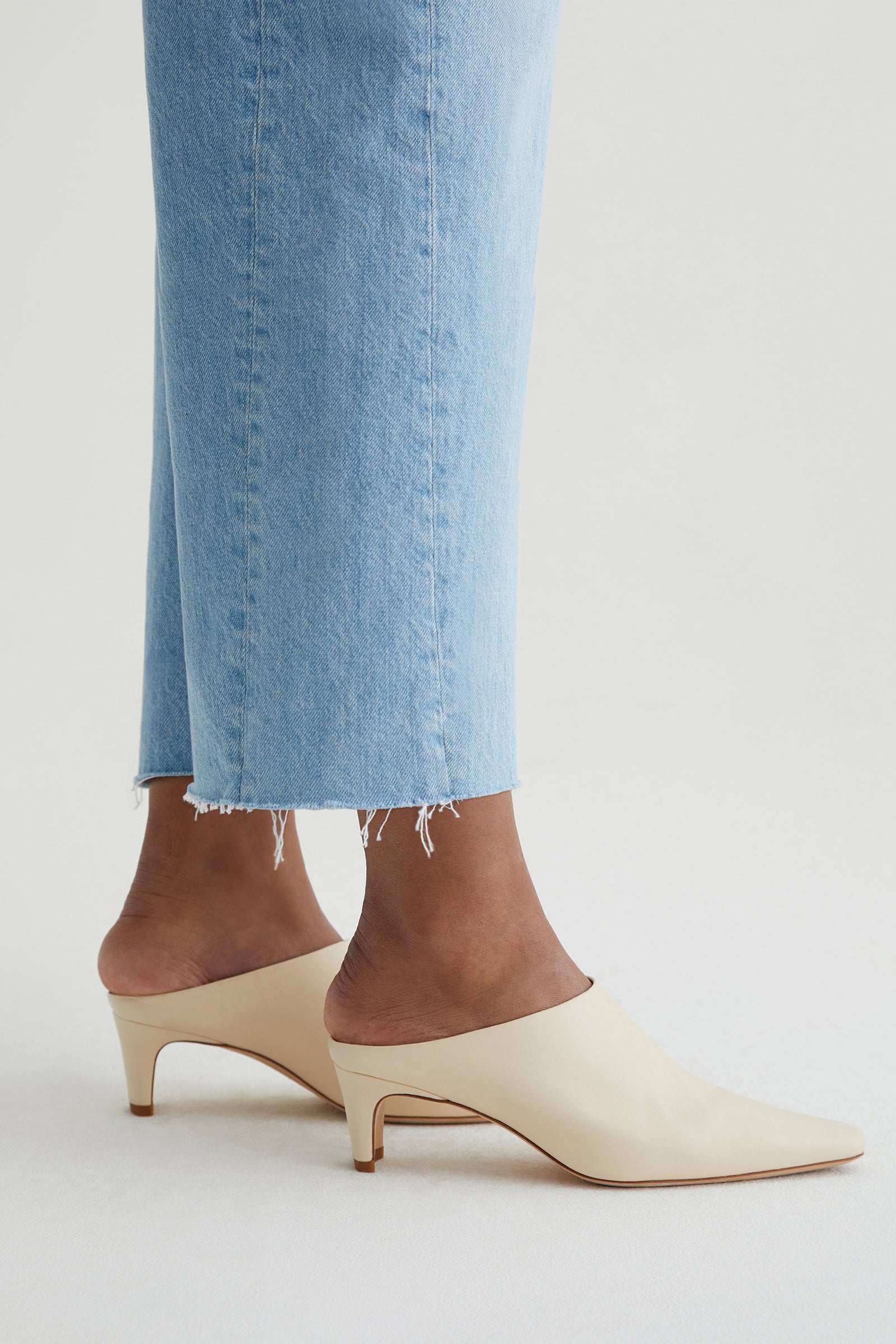Someone is wearing the AG Hattie Crop Jean in light blue, featuring a frayed hem, along with cream-colored heeled mules, while standing on a white surface.