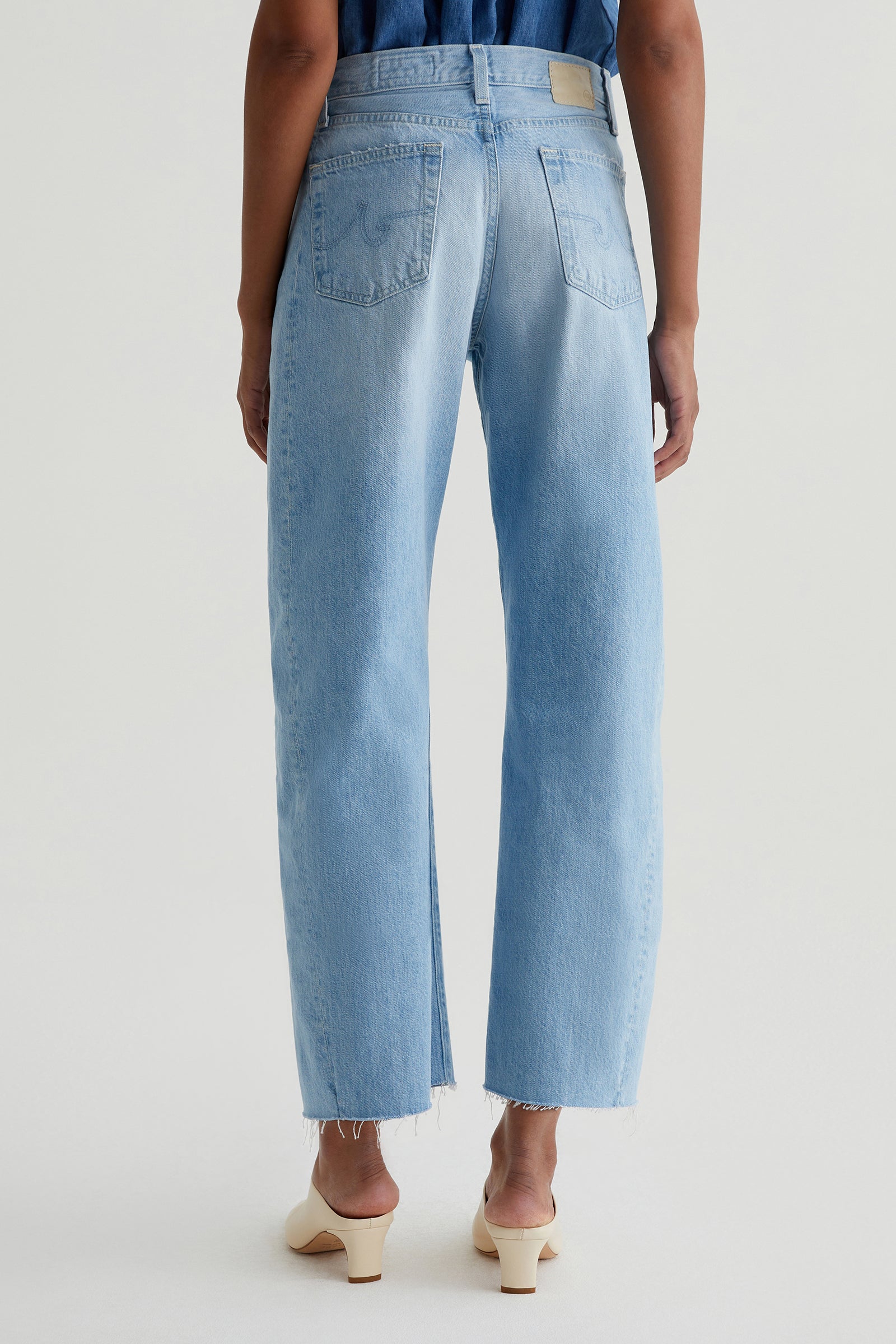 A person stands facing away, wearing the AG Hattie Crop Jean in light blue with frayed hems, paired with a sleeveless blue top and cream heeled sandals against a plain, light-colored background.