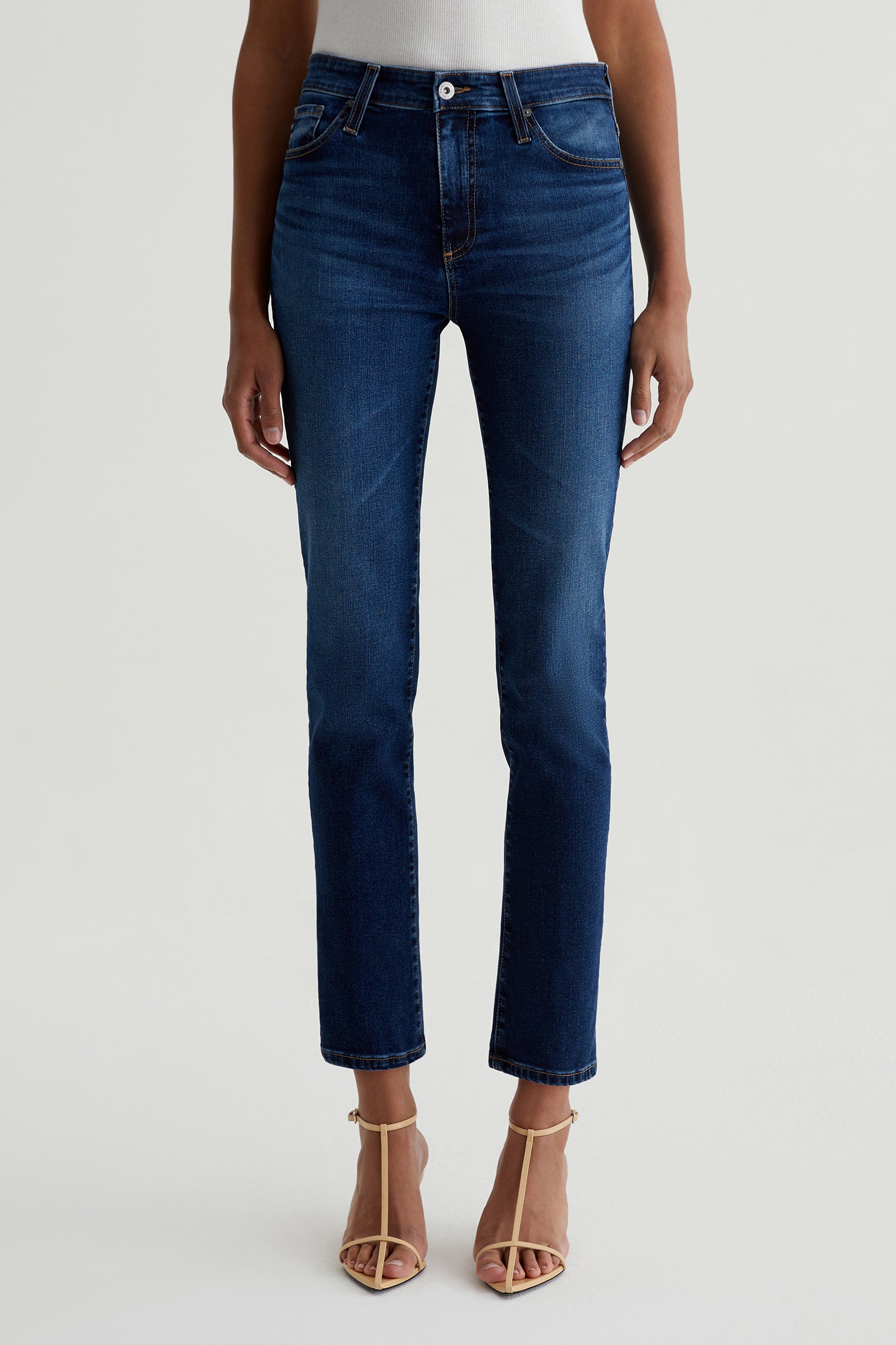 A person stands against a plain light background wearing AG Mari Slim-Straight Crop Jean in indigo with beige strappy heels and a white top tucked in; the upper body is partially visible.