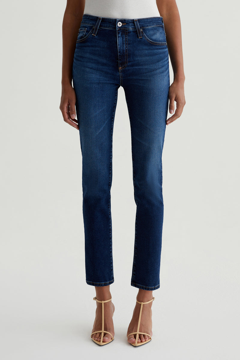 A person stands against a plain light background wearing AG Mari Slim-Straight Crop Jean in indigo with beige strappy heels and a white top tucked in; the upper body is partially visible.