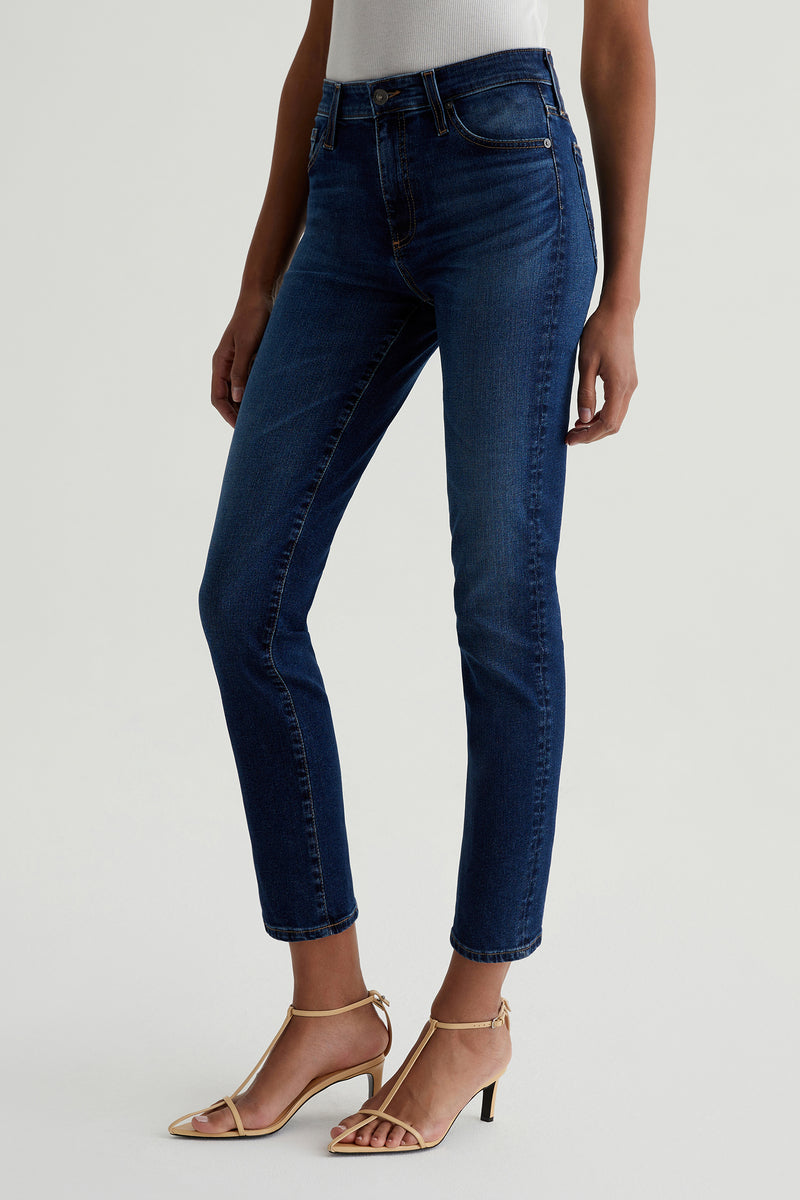 A person wearing a white top, AG Mari Slim-Straight Crop Jean - Indigo, and beige heeled sandals stands against a plain light background.