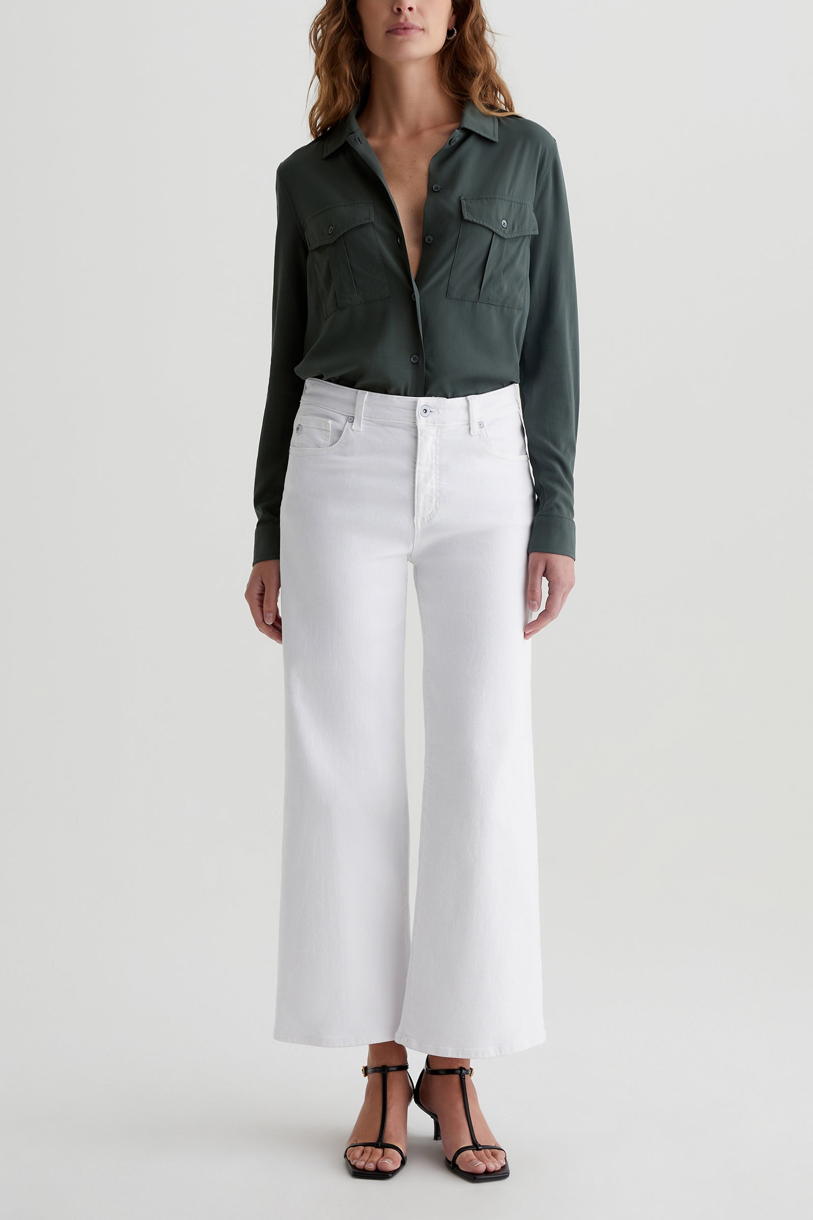 A woman stands against a plain background wearing a dark green button-up shirt, black strappy sandals, and the AG Saige Mid-Rise Wide-Leg Crop Jean in white.
