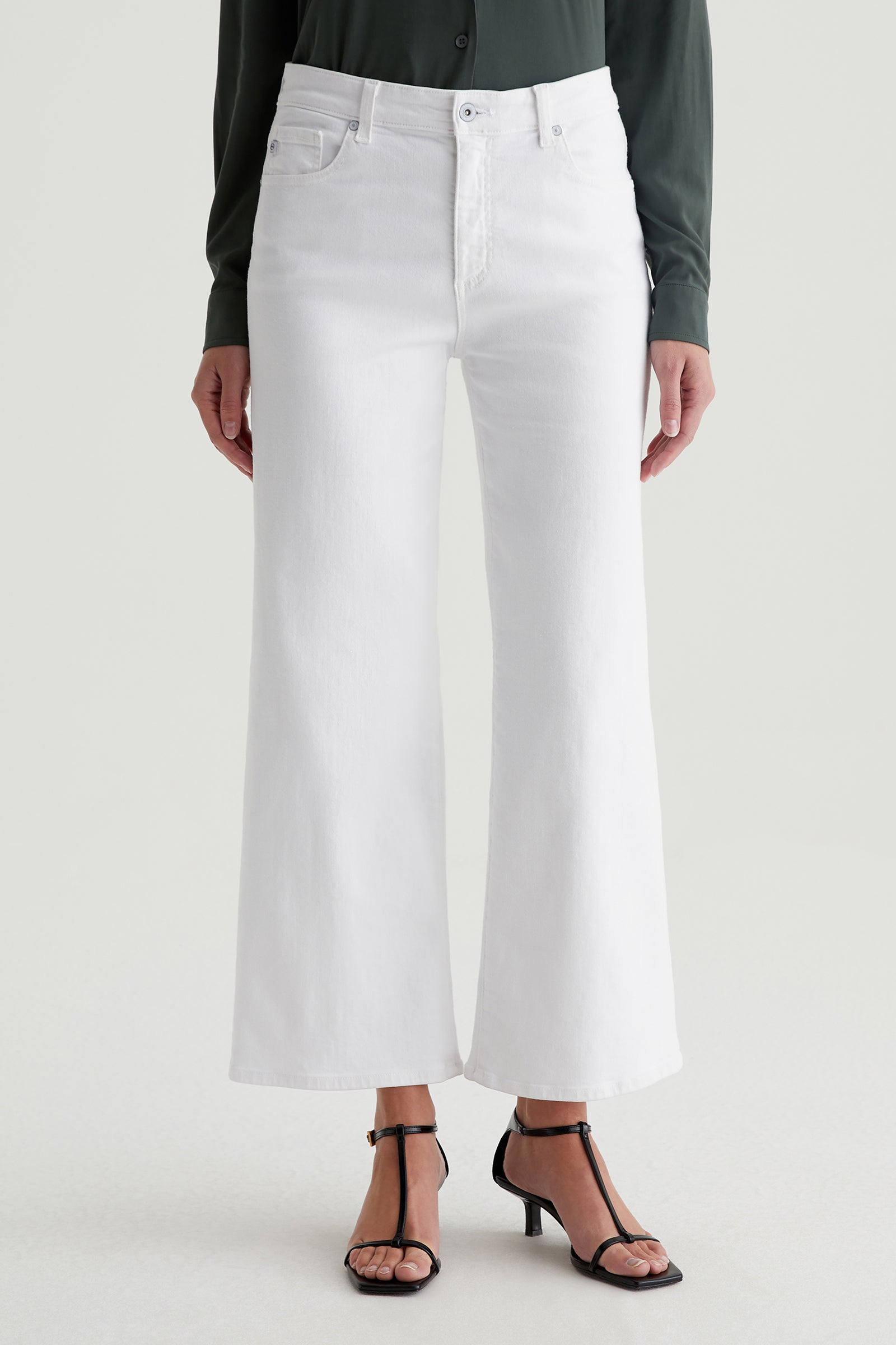 Someone wearing the AG Saige Mid-Rise Wide-Leg Crop Jean in white, paired with a dark long-sleeve shirt and black strappy heeled sandals, stands against a plain light background.