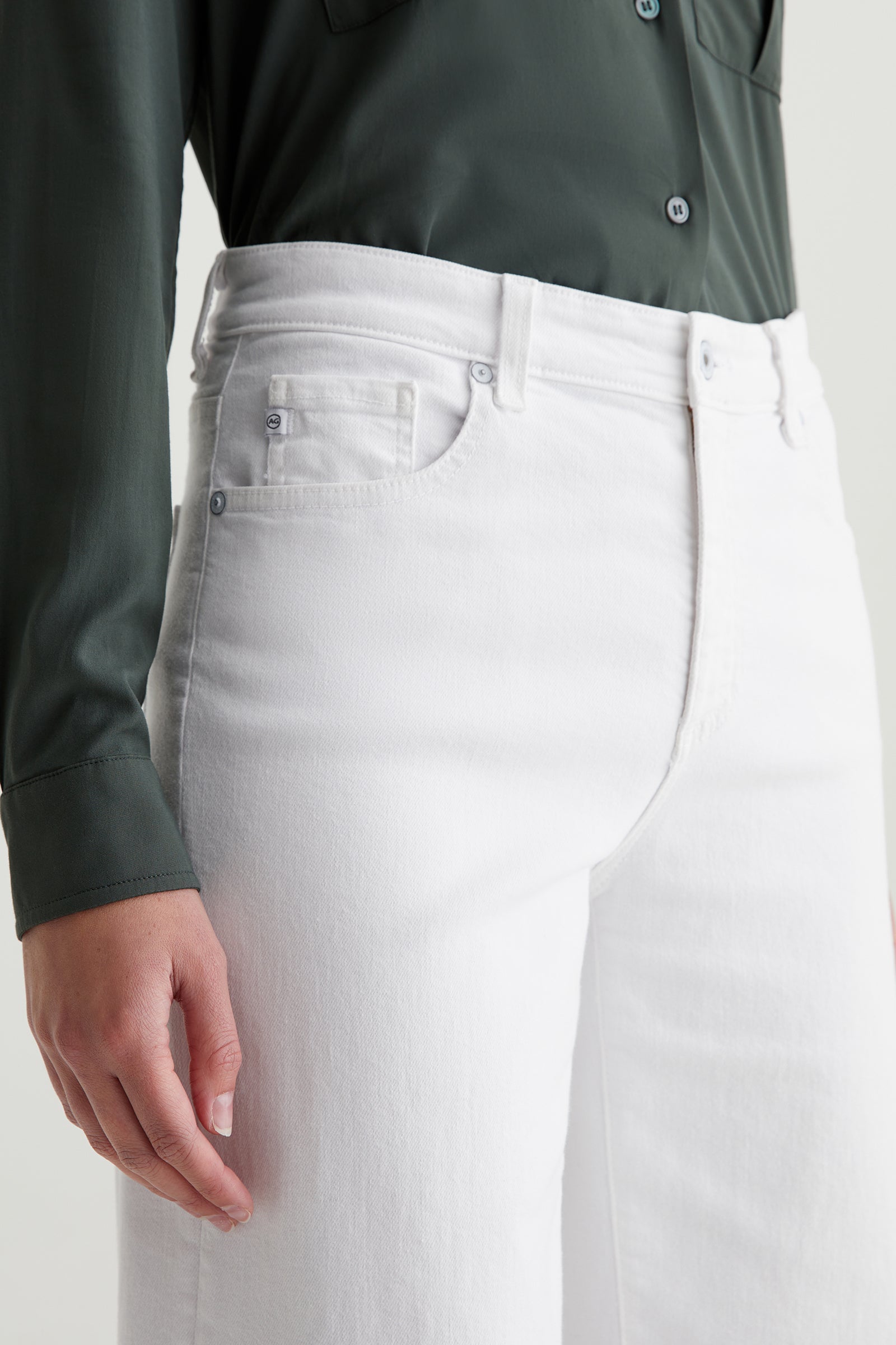 A person wearing a dark green button-up shirt and AG Saige Mid-Rise Wide-Leg Crop Jean in White, shown from mid-torso to thigh, with their left arm resting at their side.