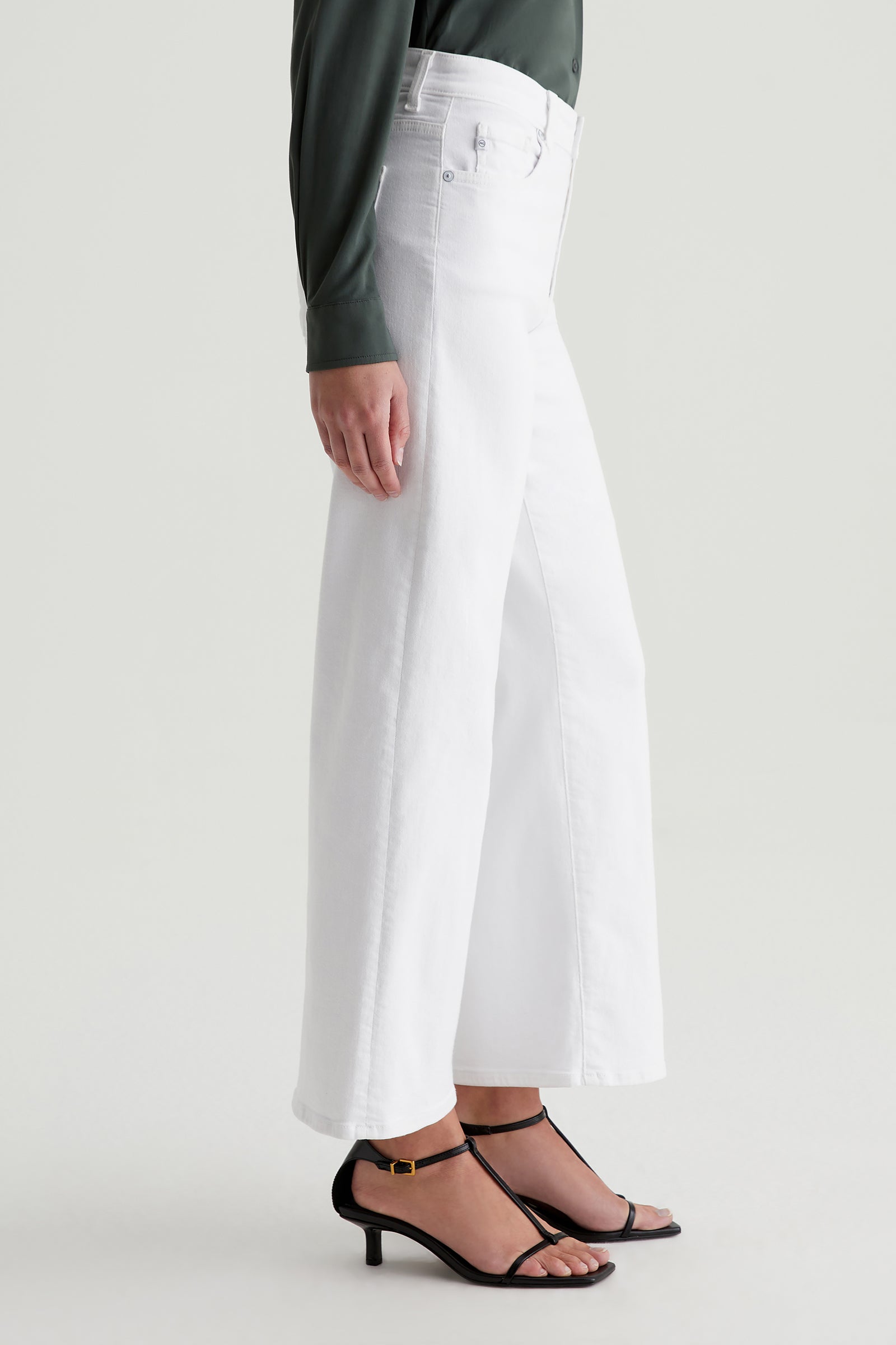A person stands sideways in AG Saige Mid-Rise Wide-Leg Crop Jeans in white, paired with a dark long-sleeve shirt and black strappy heeled sandals against a plain light background.