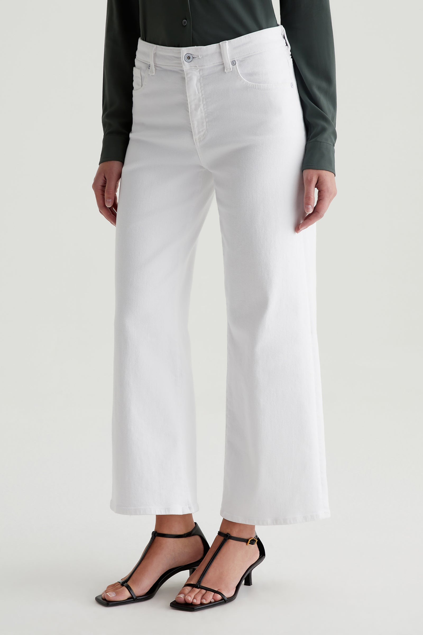A person wears AG Saige Mid-Rise Wide-Leg Crop Jeans in white, paired with a dark green long-sleeve shirt and black heeled sandals, standing against a plain light background.