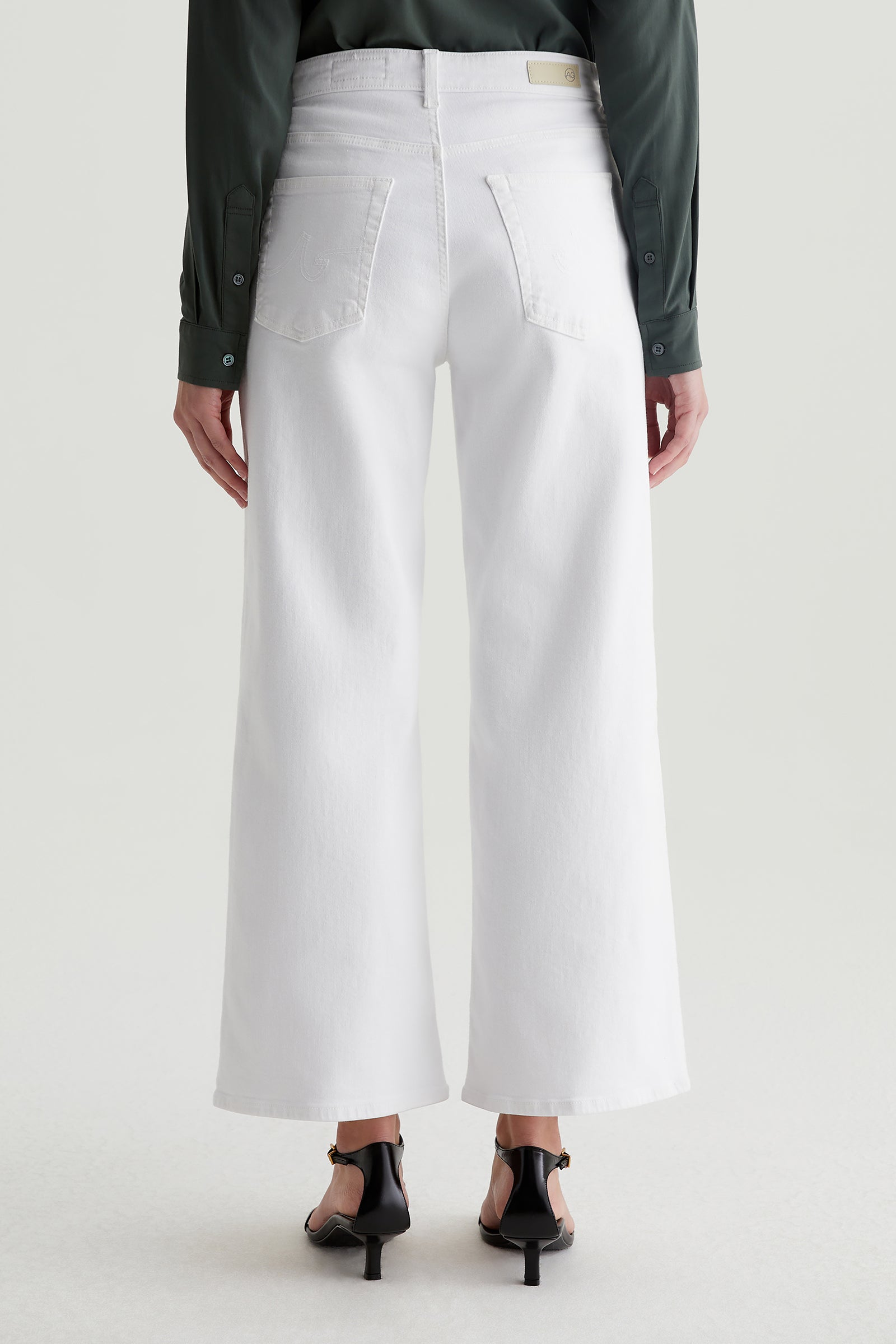 A person is seen from the back wearing an AG Saige Mid-Rise Wide-Leg Crop Jean in white, paired with a dark long-sleeve shirt and black heeled sandals, standing against a light plain background.
