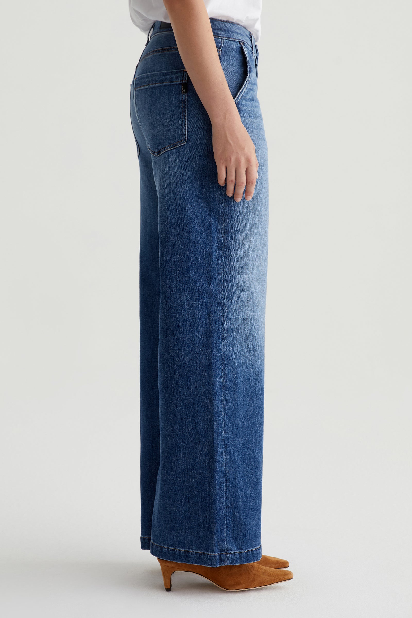 Wearing the AG Stella Low-Rise Palazzo Jean - Indigo, a person stands sideways against a plain light background, showing only the lower half of their body paired with tan heeled shoes.