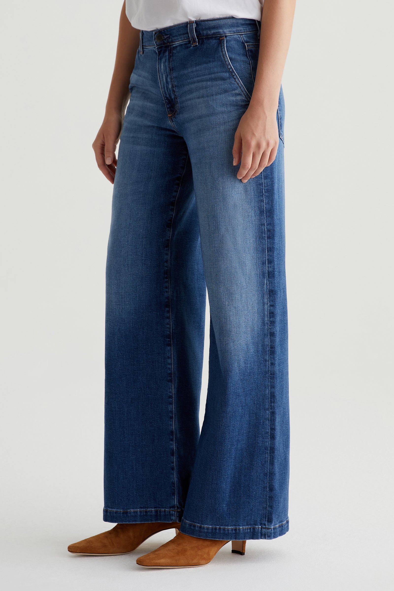 A person wearing the AG Stella Low-Rise Palazzo Jean in indigo, paired with a white top and tan heeled boots, stands against a plain light background.