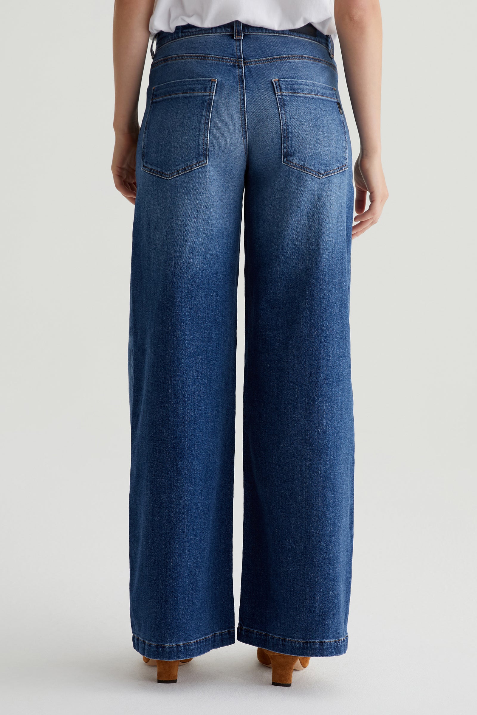 A person stands facing away from the camera in a white top and AG Stella Low-Rise Palazzo Jean - Indigo, featuring back pockets, wide legs, a slight faded effect, and brown-heeled shoes.