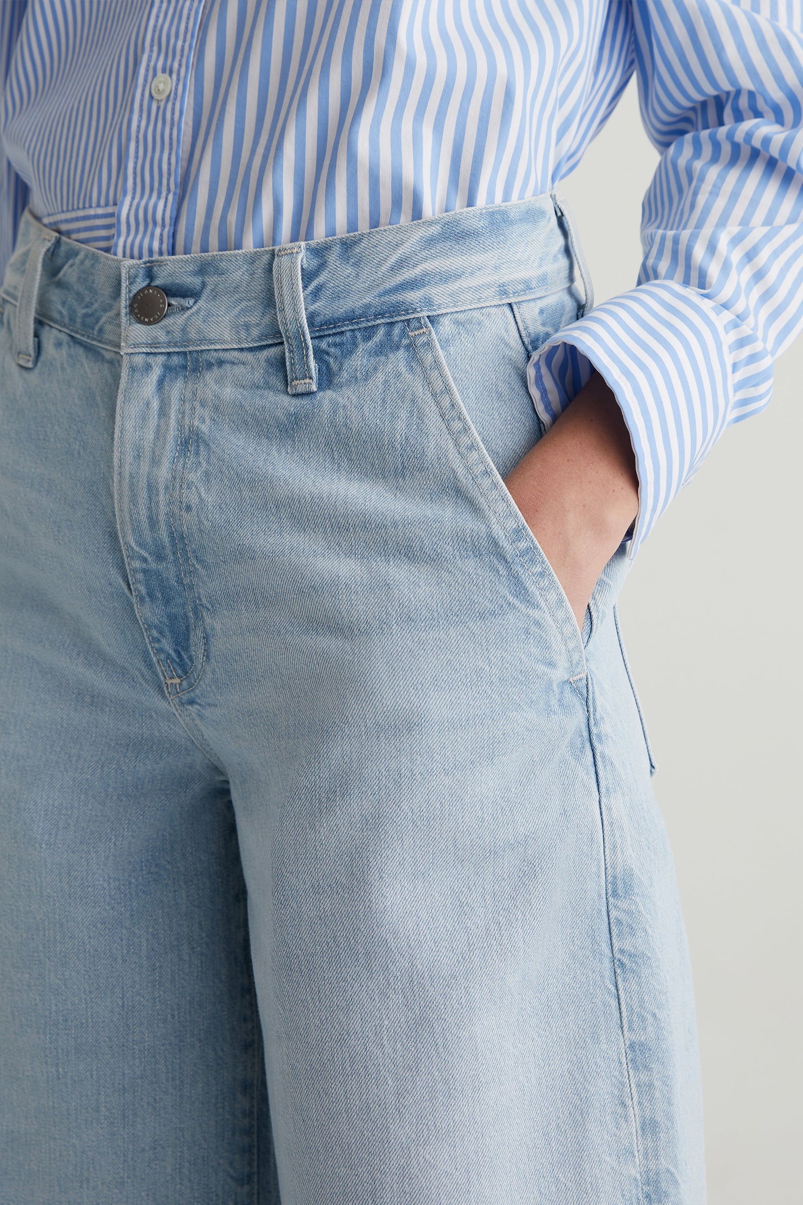 A person wears a blue and white striped button-up shirt with their hand in the pocket of AG Stella Low-Rise Palazzo Jean in light blue.