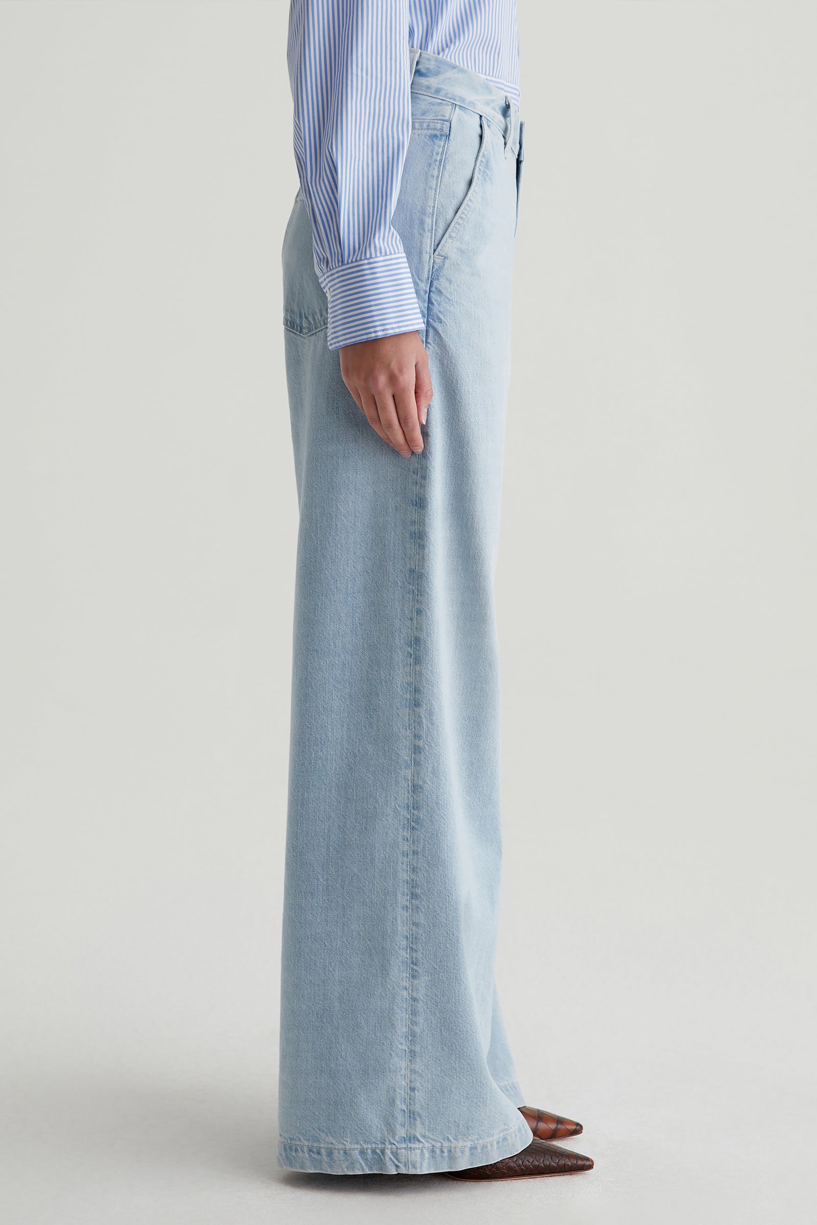 Person standing sideways in AG Stella Low-Rise Palazzo Jean - Light Blue, a blue and white striped long-sleeve shirt, and brown pointed-toe shoes, set against a plain light background.