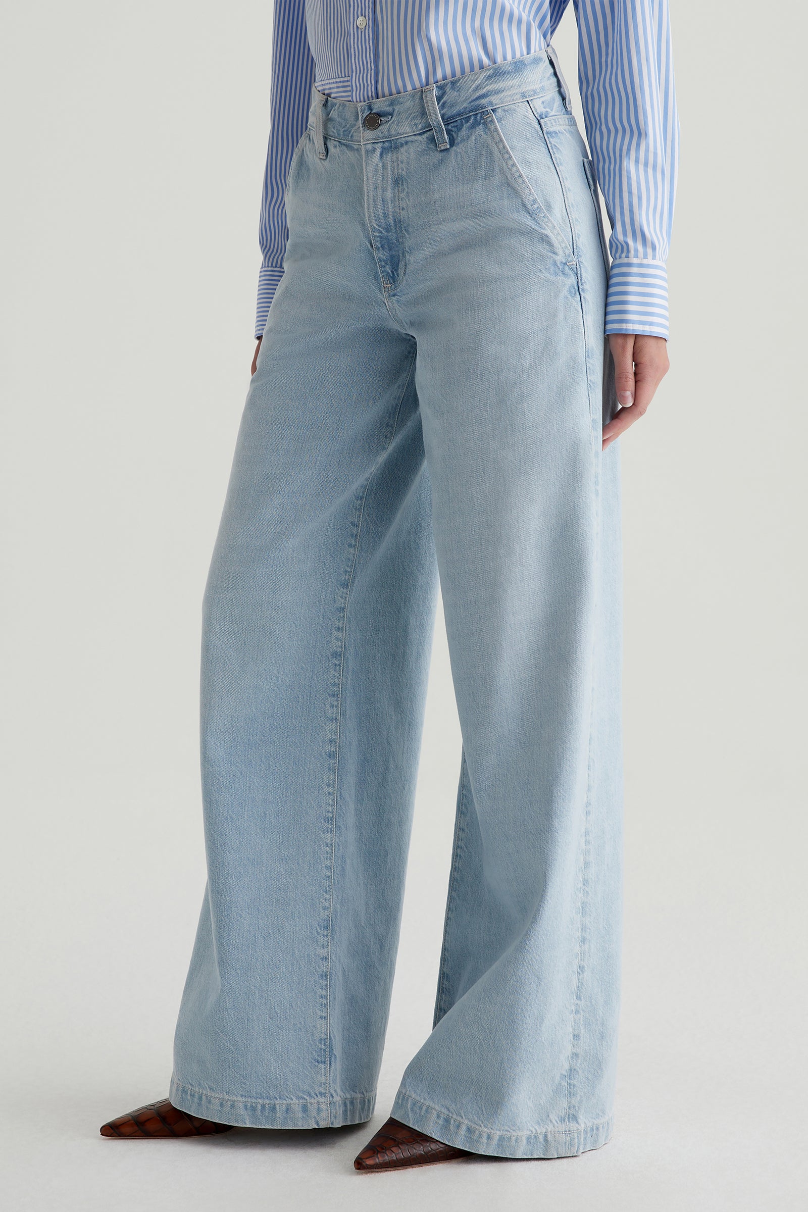 A person wearing the AG Stella Low-Rise Palazzo Jean in light blue, paired with a blue and white striped button-up shirt and pointed brown shoes, stands against a plain light background.