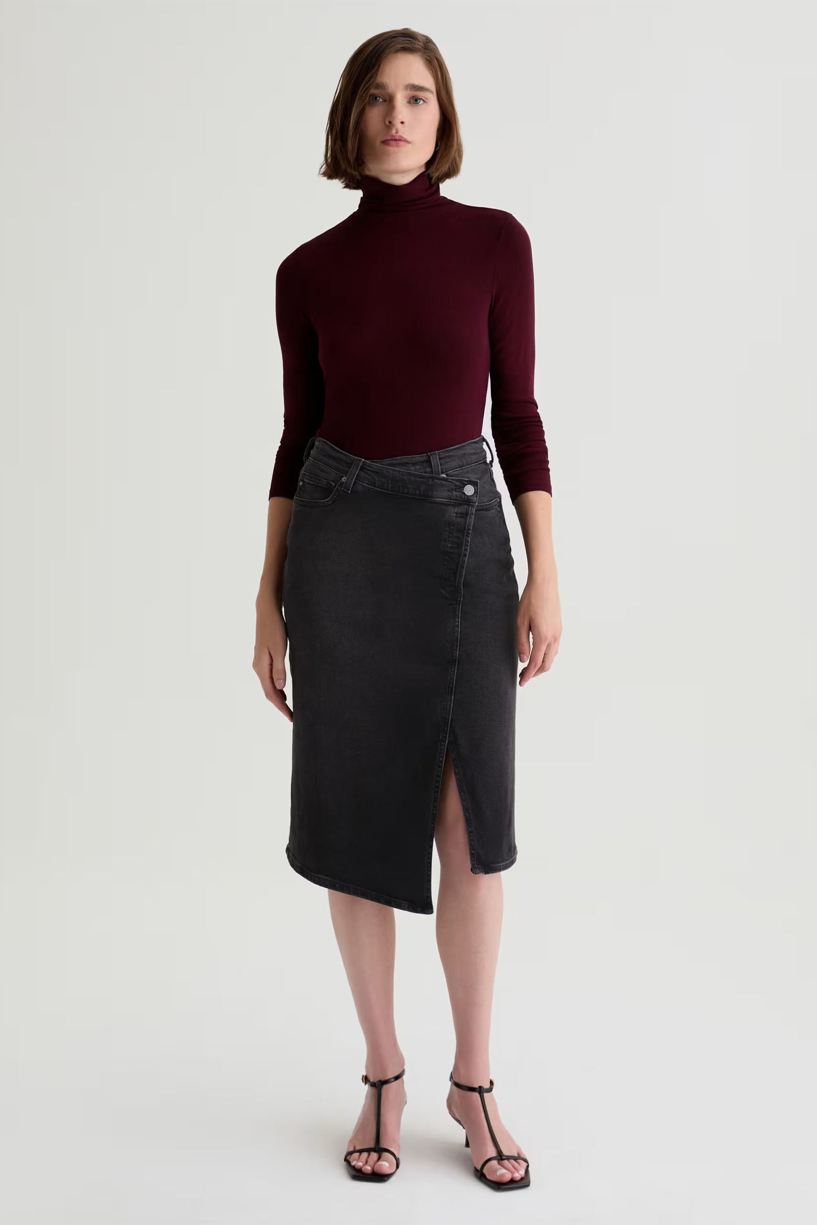 A person wears a burgundy turtleneck, the Bruna Wrap Midi Skirt in black with a front slit, and black heeled sandals against a plain light background.